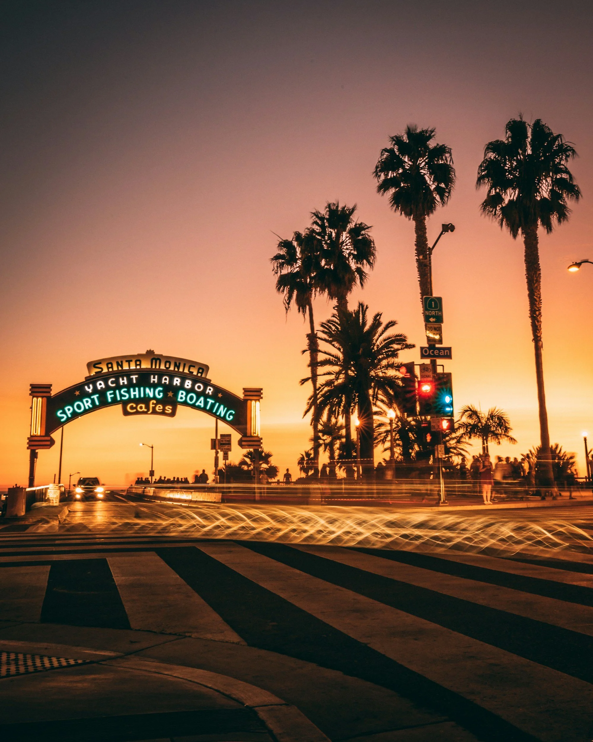 Sunset over Santa Monica Pier with tall palm trees, traffic lights, and cars passing by, creating blurred light streaks in the foreground.