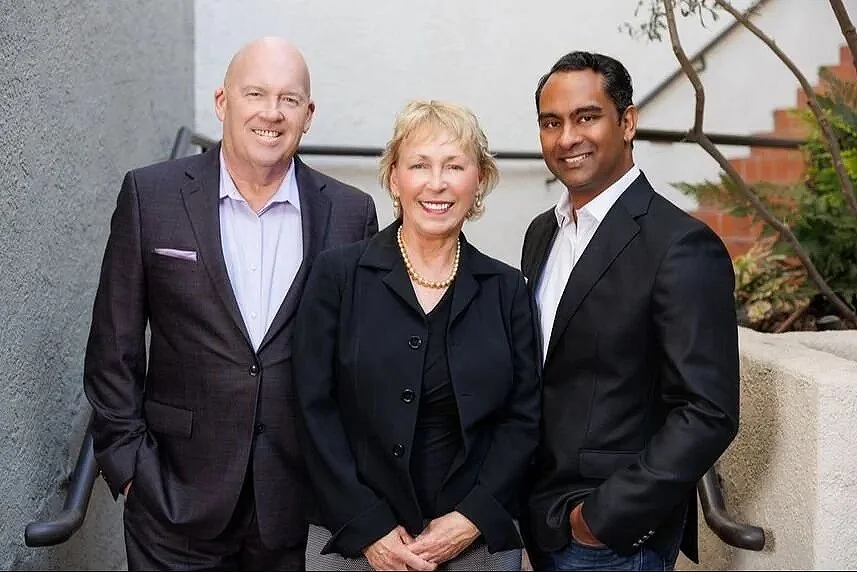 Three professionally dressed individuals smiling outdoors, two men and one woman, standing on stairs with a gray wall and some trees in the background.
