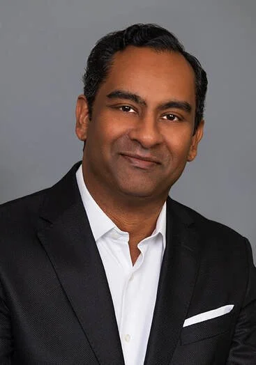 Professional headshot of a man in a black suit and white shirt with a gray background.