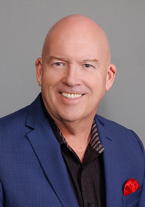 A smiling middle-aged man with a bald head, wearing a navy blue blazer, black shirt, and a red pocket square, against a gray background.