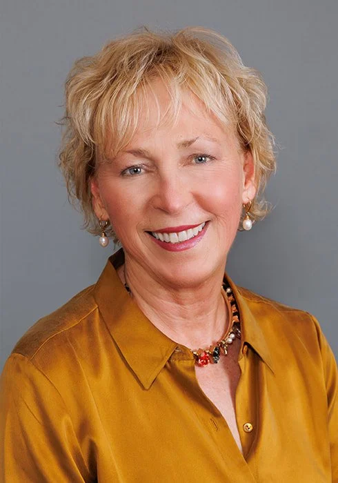Portrait of a smiling woman with short blonde hair, wearing pearl earrings, a necklace, and a mustard-colored blouse against a gray background.