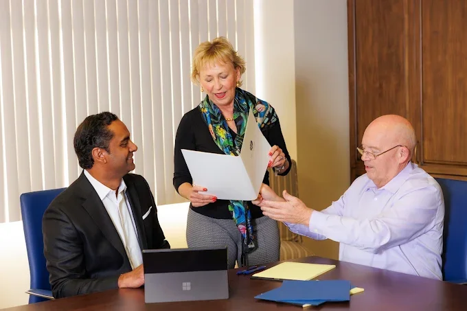 Three business professionals in an office meeting, with a woman handing a document to an older man, while a younger man looks on and a laptop is on the table.