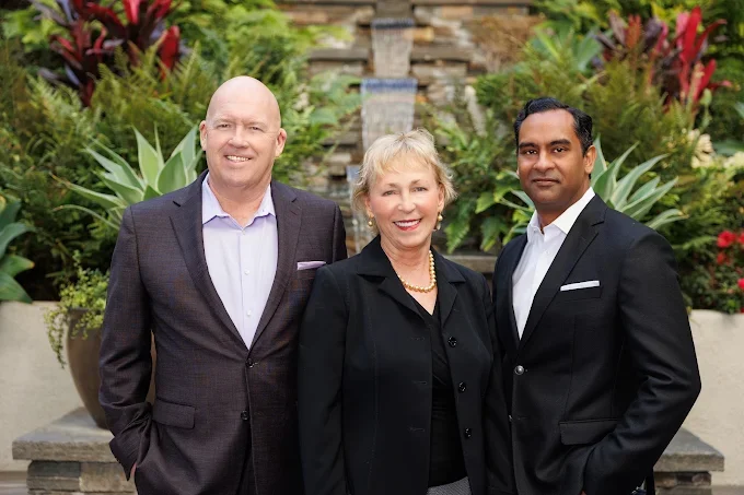 Three professionally dressed people standing outdoors in front of a garden with lush plants and a water feature, smiling at the camera.