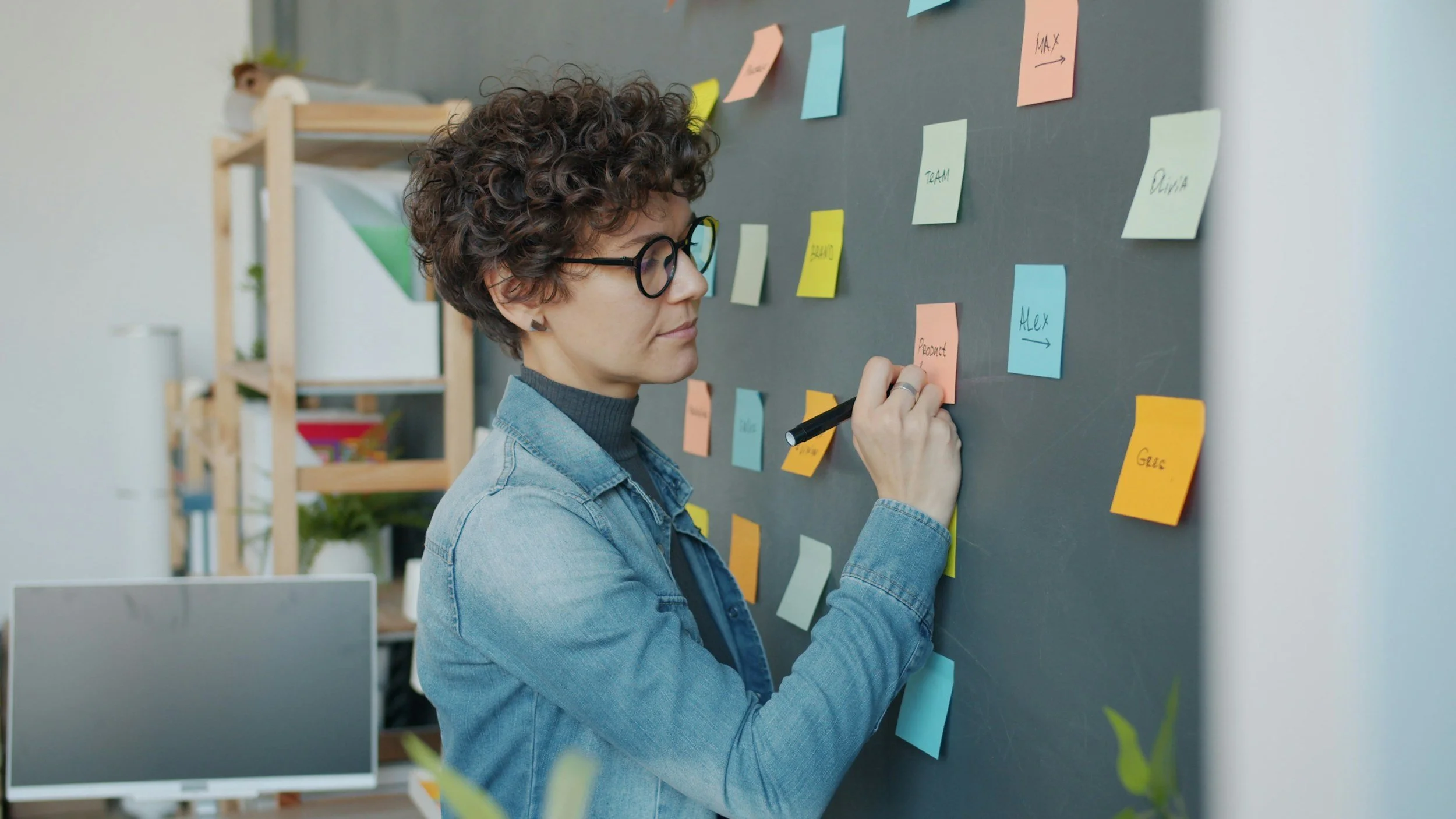 A woman wearing glasses and a denim jacket writing on a pink sticky note on a blackboard covered with various colorful sticky notes in a workspace.