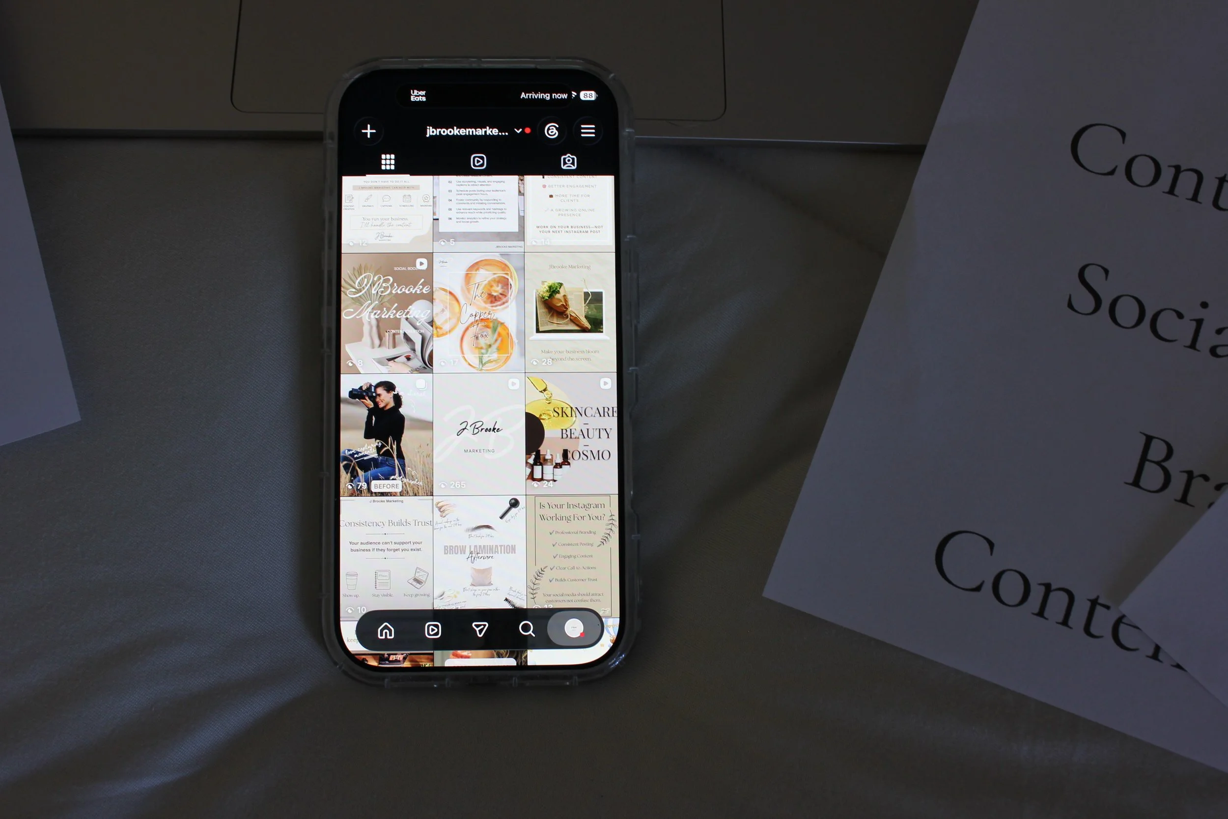 Smartphone displaying an Instagram profile with various posts, placed on a dark surface beside a white paper with partially visible black text.
