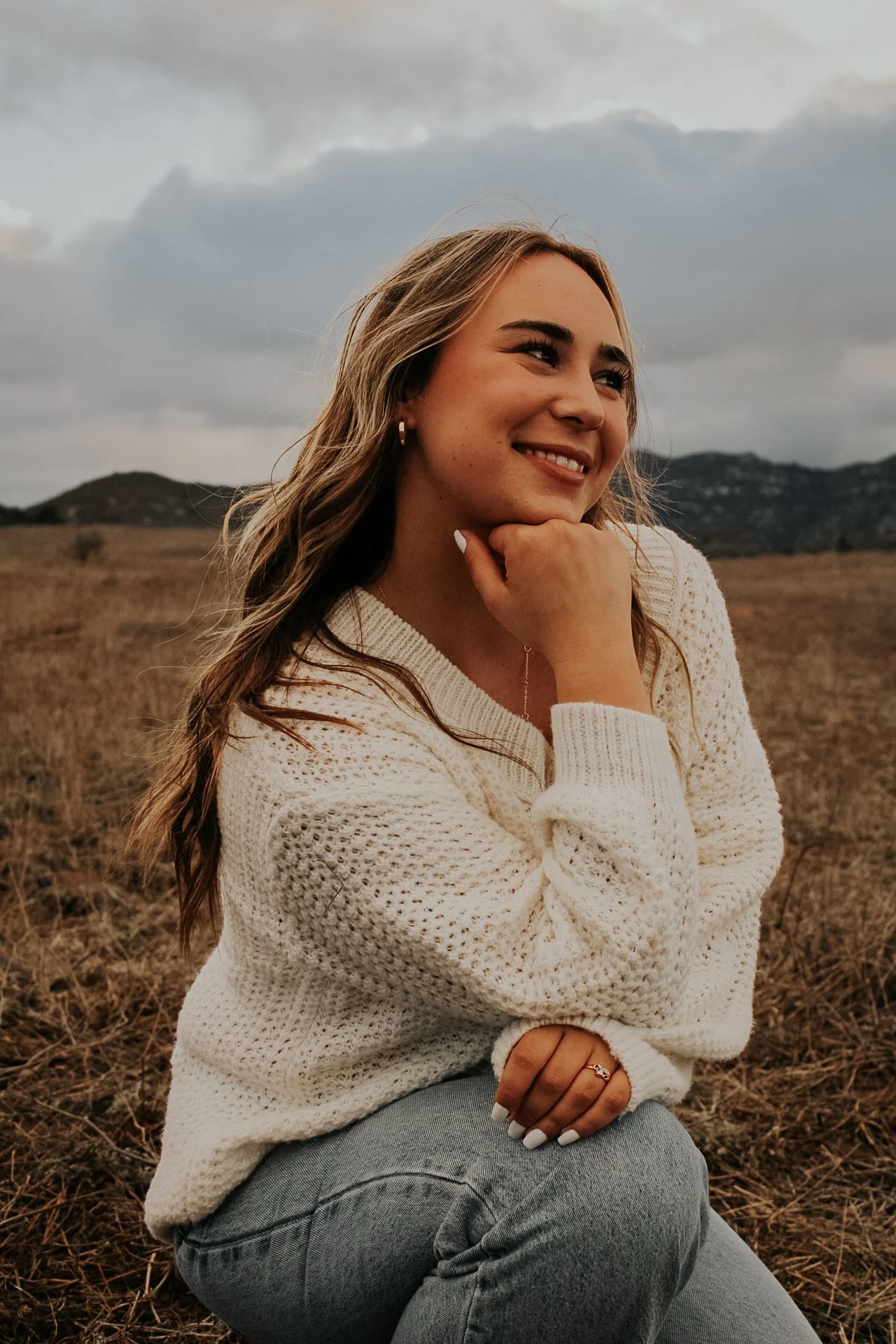 A woman sitting outdoors in a field with mountains in the background, smiling and looking to the side, wearing a cream knit sweater and gray jeans.