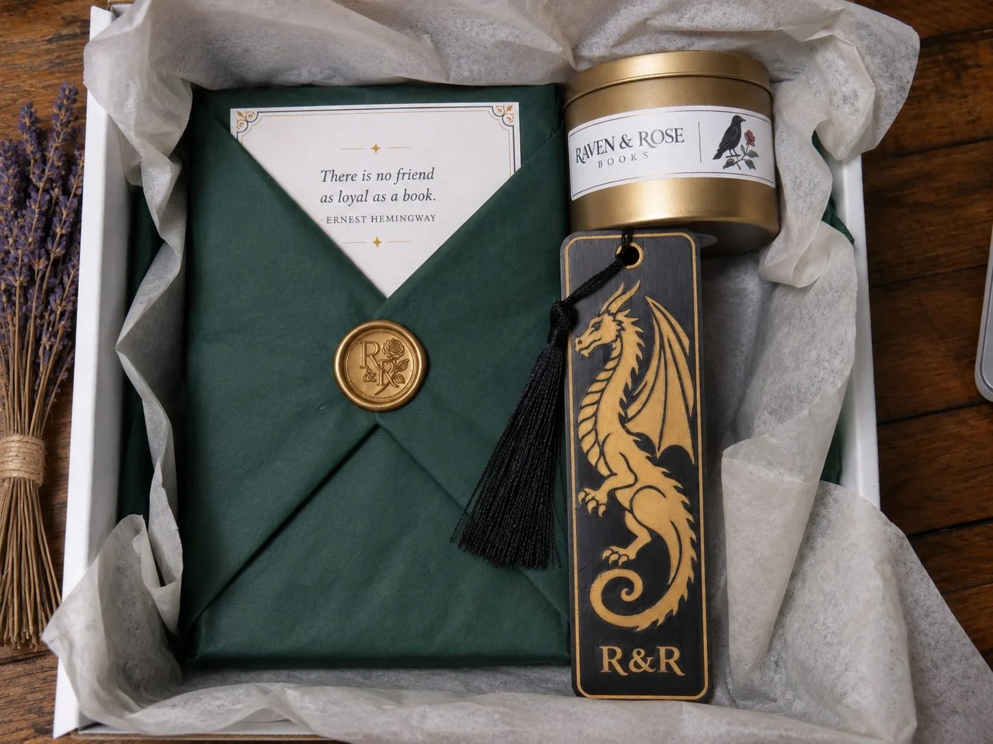 A gift box containing a sealed green envelope with a gold wax seal, a quote card from Ernest Hemingway, a gold tin labeled Raven & Rose Books, and a rectangular bookmark with a black and gold dragon design and a black tassel. The box is lined with tissue paper and placed on a wooden surface, with a bunch of dried lavender on the side.