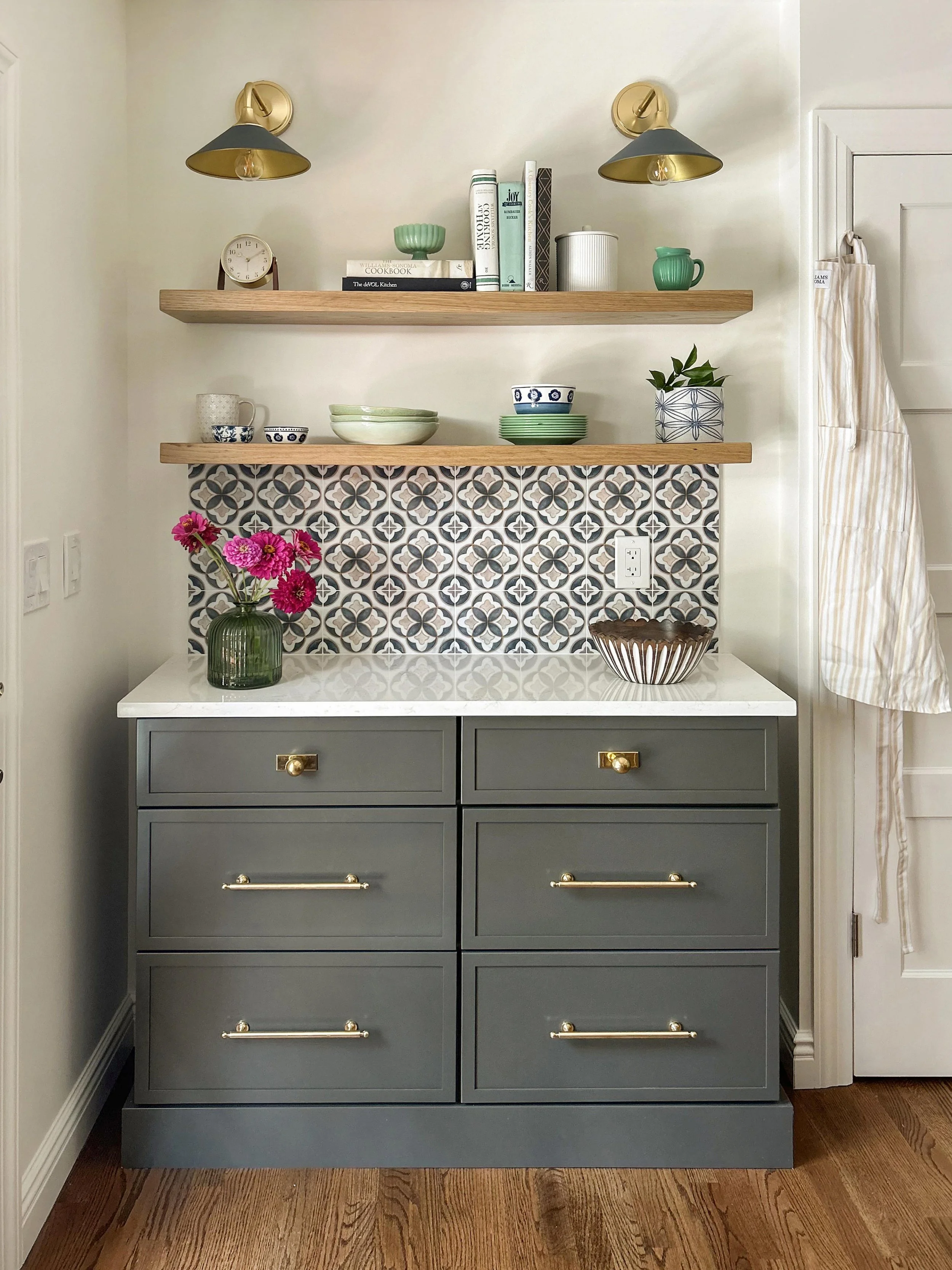 A grey kitchen cabinet with four drawers, gold handles, a white countertop, and a patterned tile backsplash. There are two wooden shelves above the cabinet holding various bowls, cups, books, and decorative items. A pink flower bouquet in a green vase is on the countertop. Two wall-mounted lamps with grey and gold shades are above the shelves, and a folded striped cloth is hanging on a door to the right.