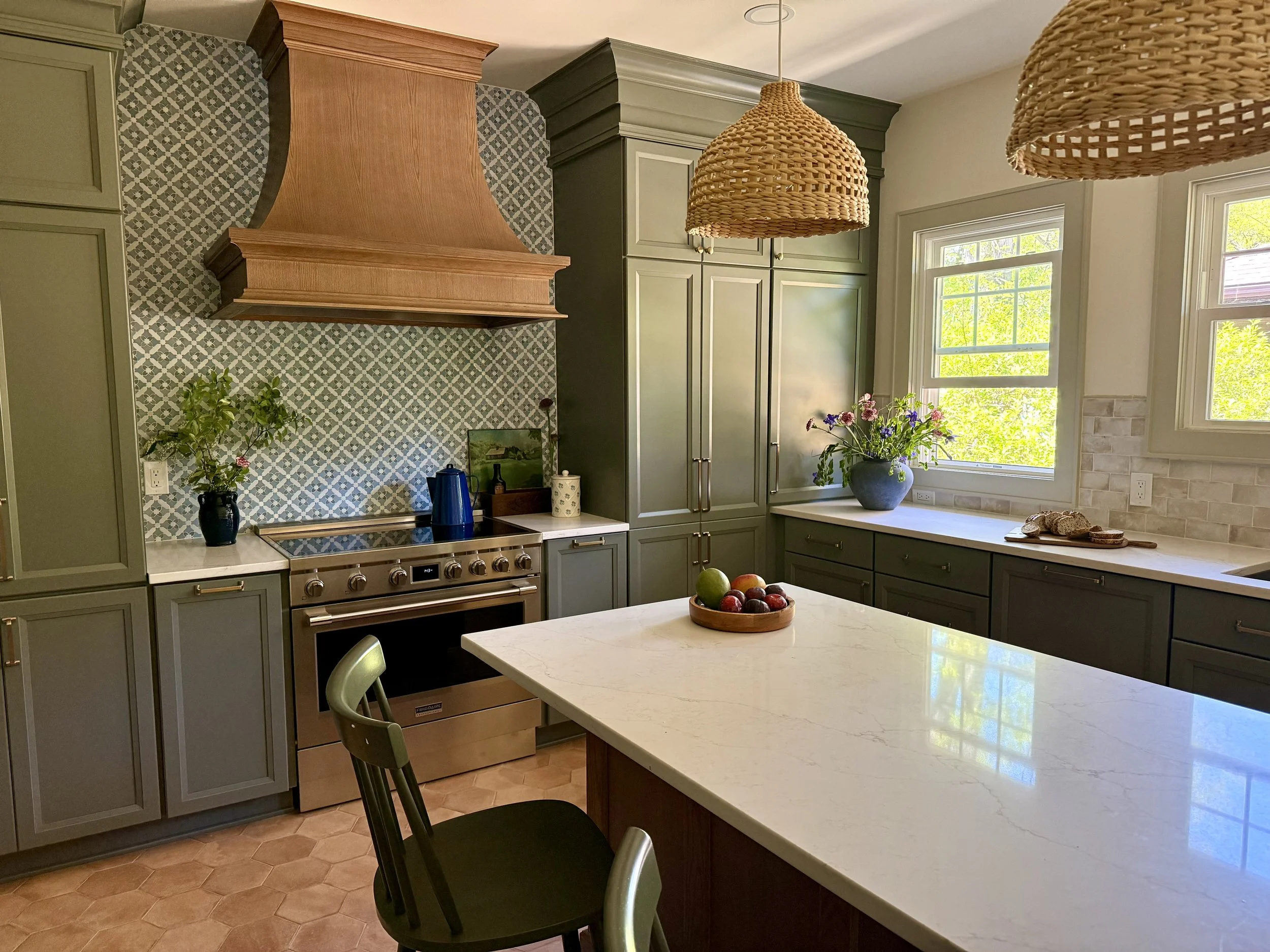 Kitchen with green cabinets, a black range hood, a white marble countertop, and a large window with green foliage outside. There are two woven pendant lights hanging from the ceiling, a bowl of apples on the island, and decorative plants on the counter.