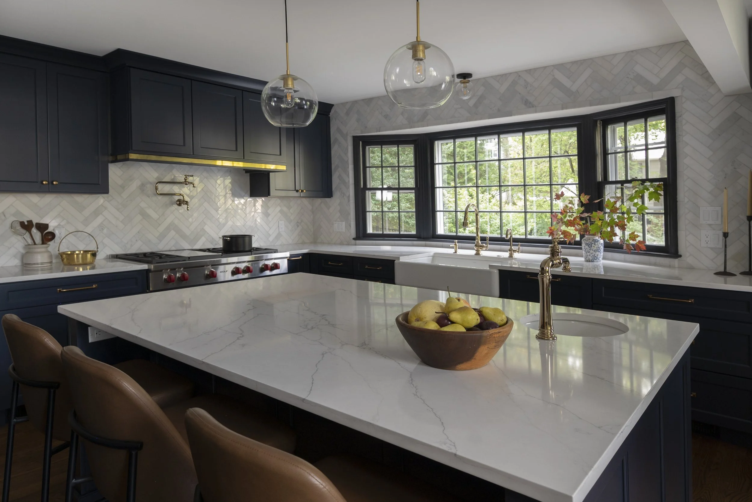 Modern kitchen with black cabinets, white marble countertops, a large window, gold fixtures, and a bowl of apples on the island.