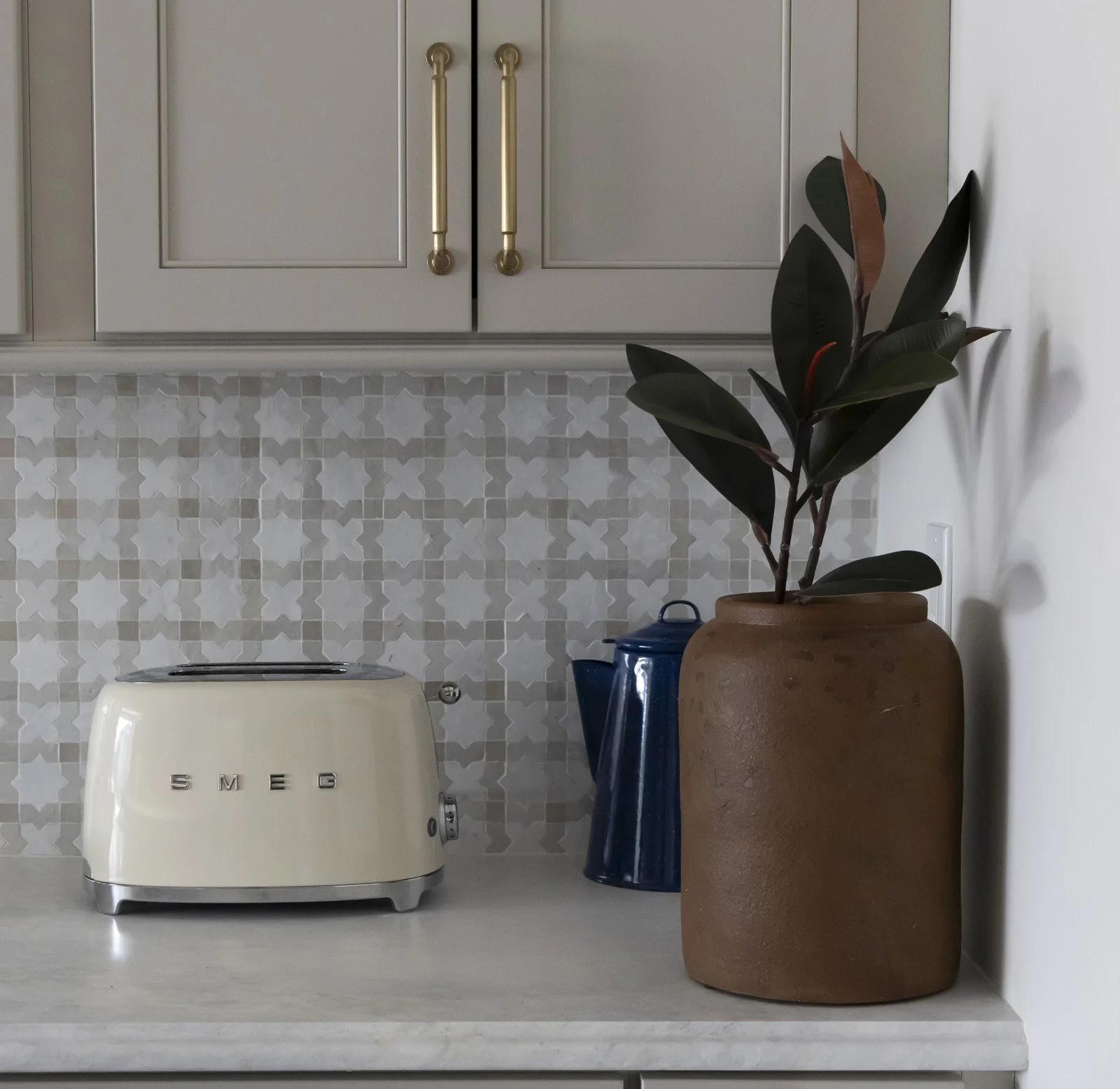 A kitchen counter in one of the kitchens designed by Julie Sidwell Design, faturing a cream-colored SMEG toaster, a large terracotta pot with a plant, a blue ceramic teapot, and a patterned backsplash. Cabinets with gold handles are above.