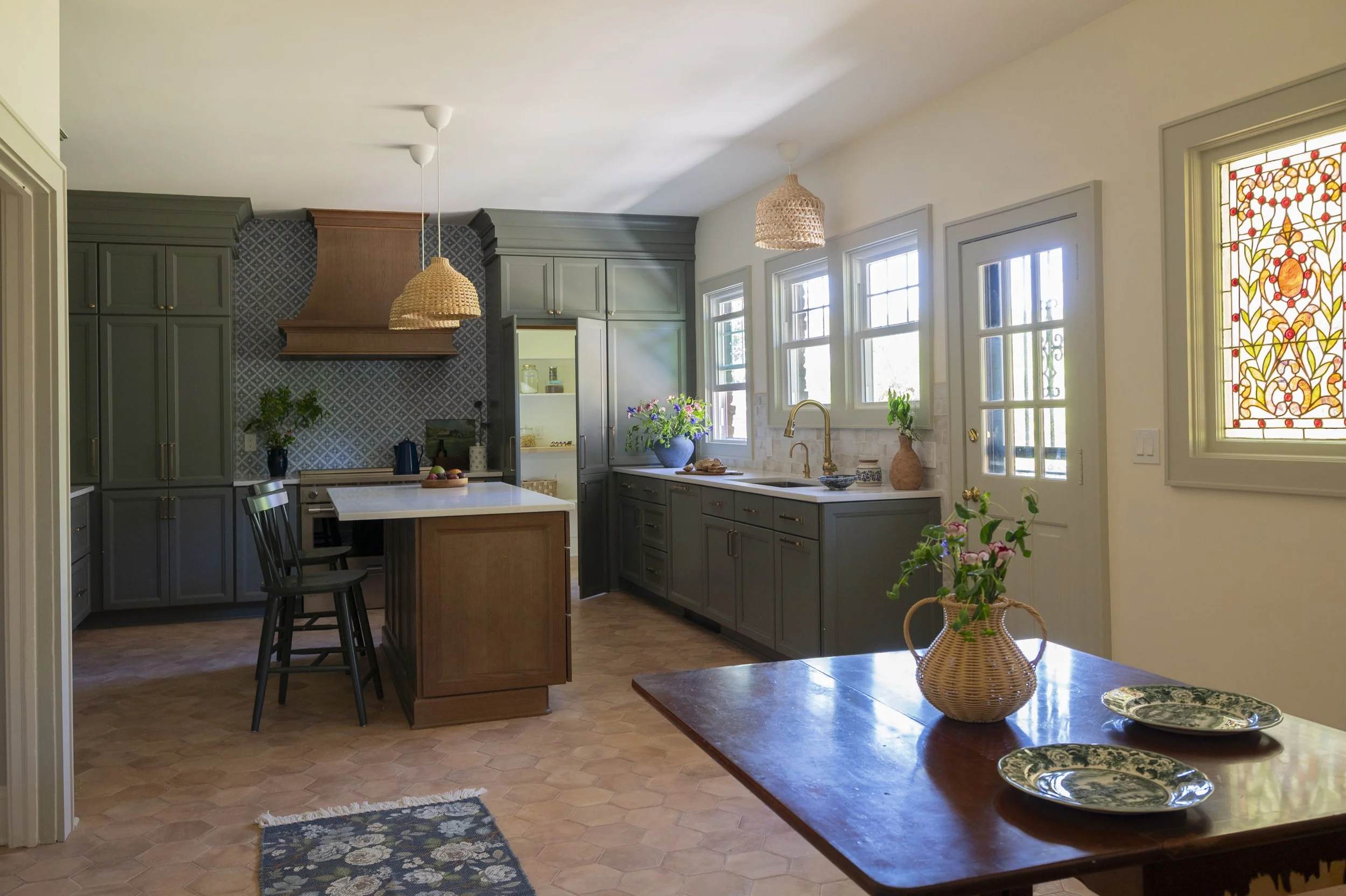 A bright farmhouse kitchen with green cabinets, a white island, and a wooden dining table with decorative plates. Features large windows, a stained-glass window on the right, and woven pendant lights hanging from the ceiling.