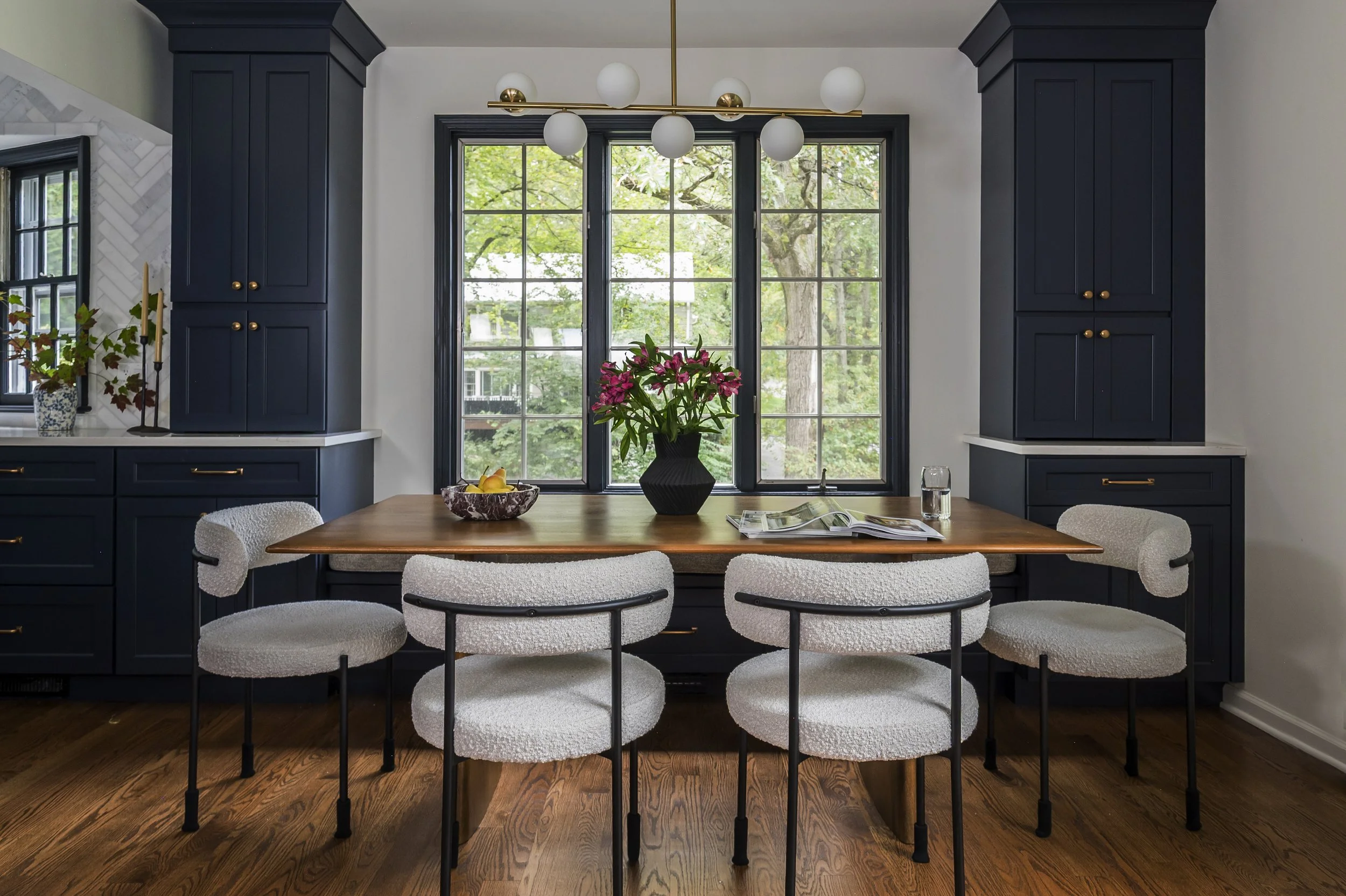 A kitchen dining area with a wooden table surrounded by four white, cushioned chairs with black metal frames. A black vase with pink flowers is on the table, along with some bowls of fruit, a glass of water, and magazines. Behind the table, large multi-pane windows show green trees outside. Dark blue cabinets and a modern gold and white globe pendant light are also visible.