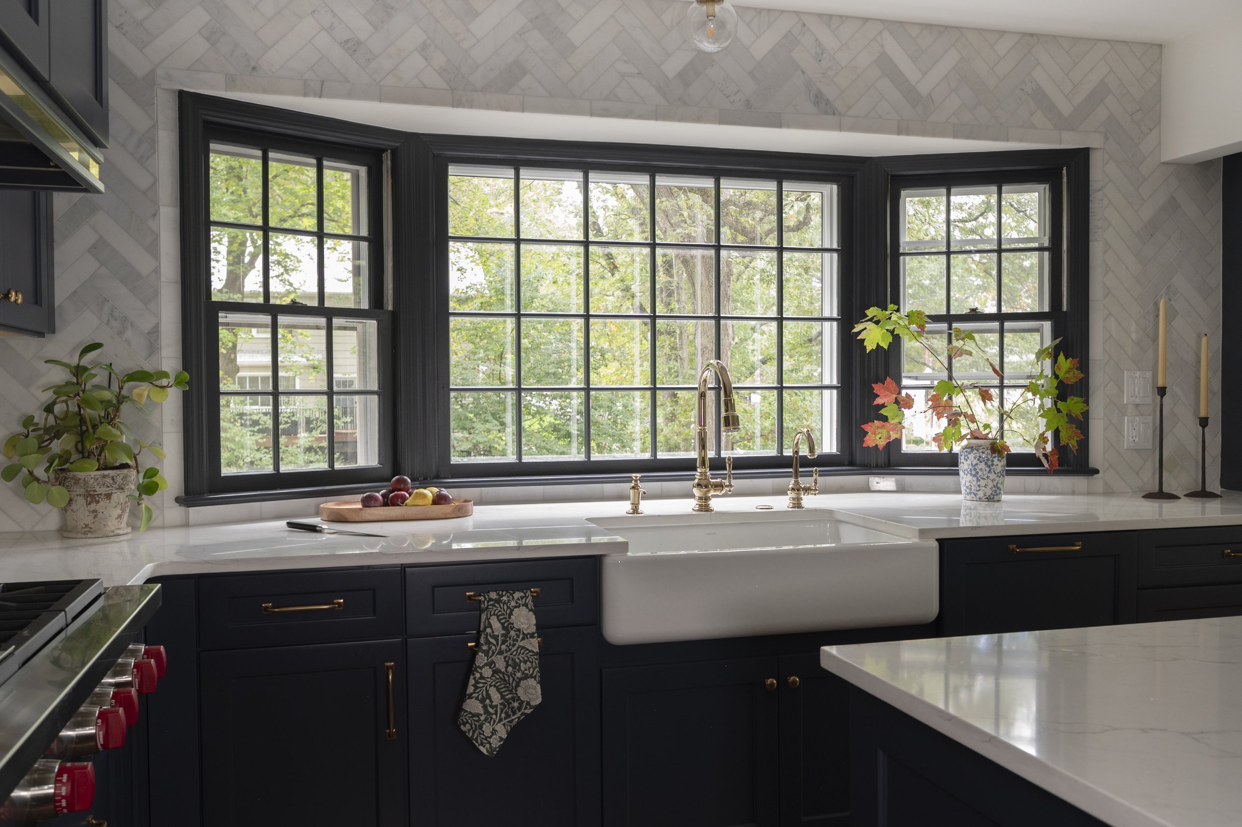 A kitchen with black window frames, white countertops, and dark blue cabinets. There is a large window above the sink showing green trees outside. Decor includes a potted plant, a bowl of apples and pears, and a blue and white vase with a branch. The backsplash features a herringbone tile pattern.