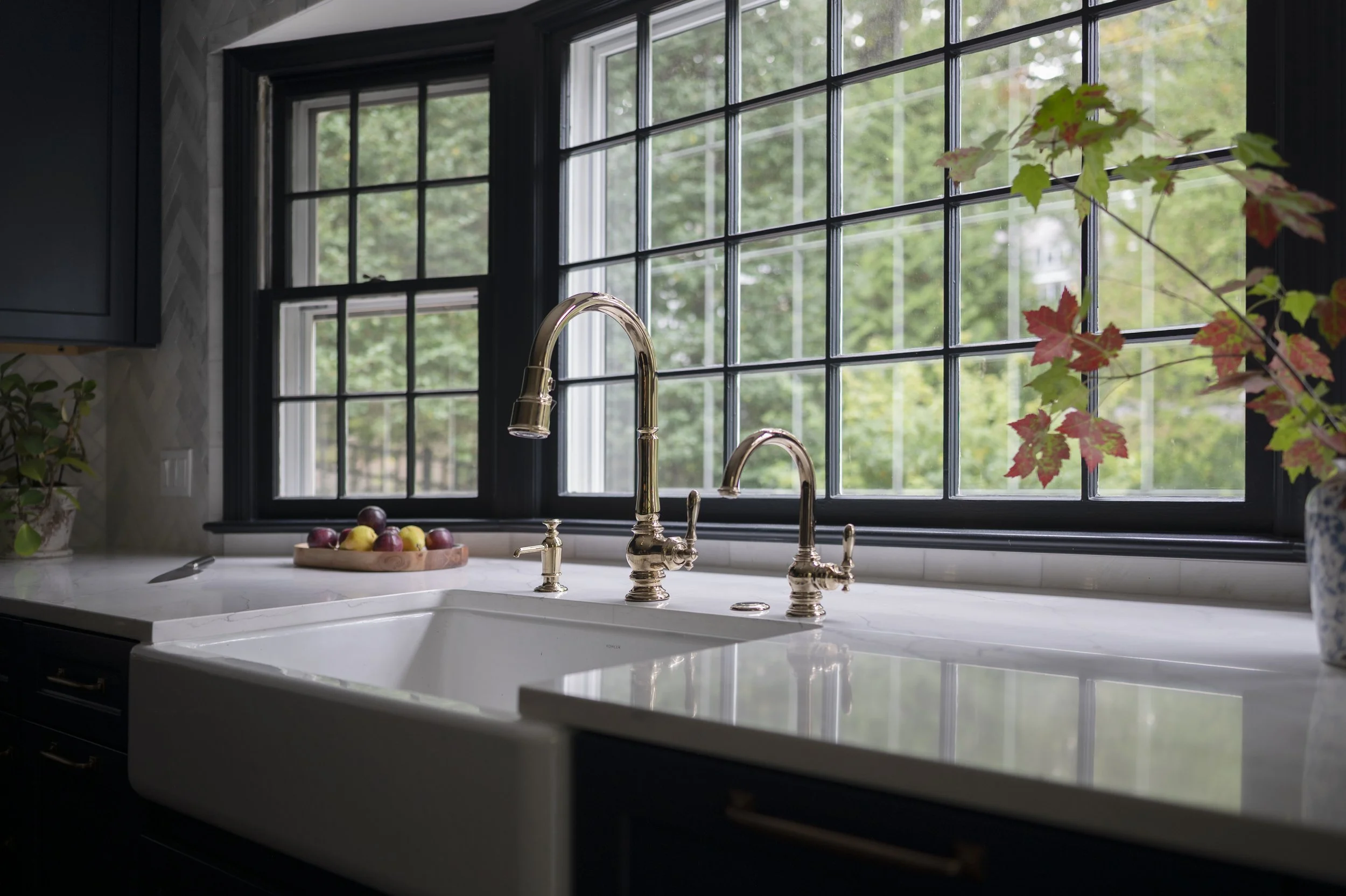 A kitchen window with black grid framing, a marble countertop, gold faucet, a bowl of assorted fruits, and greenery outside.