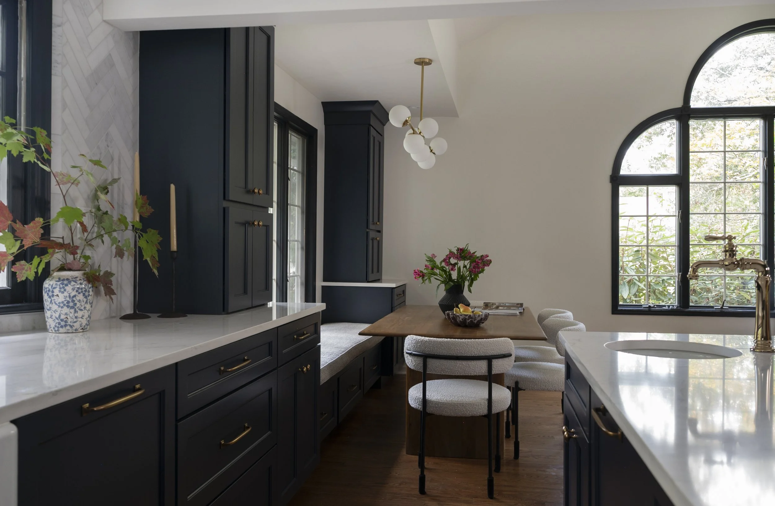 A modern kitchen with navy blue cabinets, white marble countertops, and a wooden dining table with cushioned chairs. Large arched windows bring in natural light, and a contemporary chandelier hangs from the ceiling. Decor includes a vase with flowers, a bowl of fruit, and a book on the table.