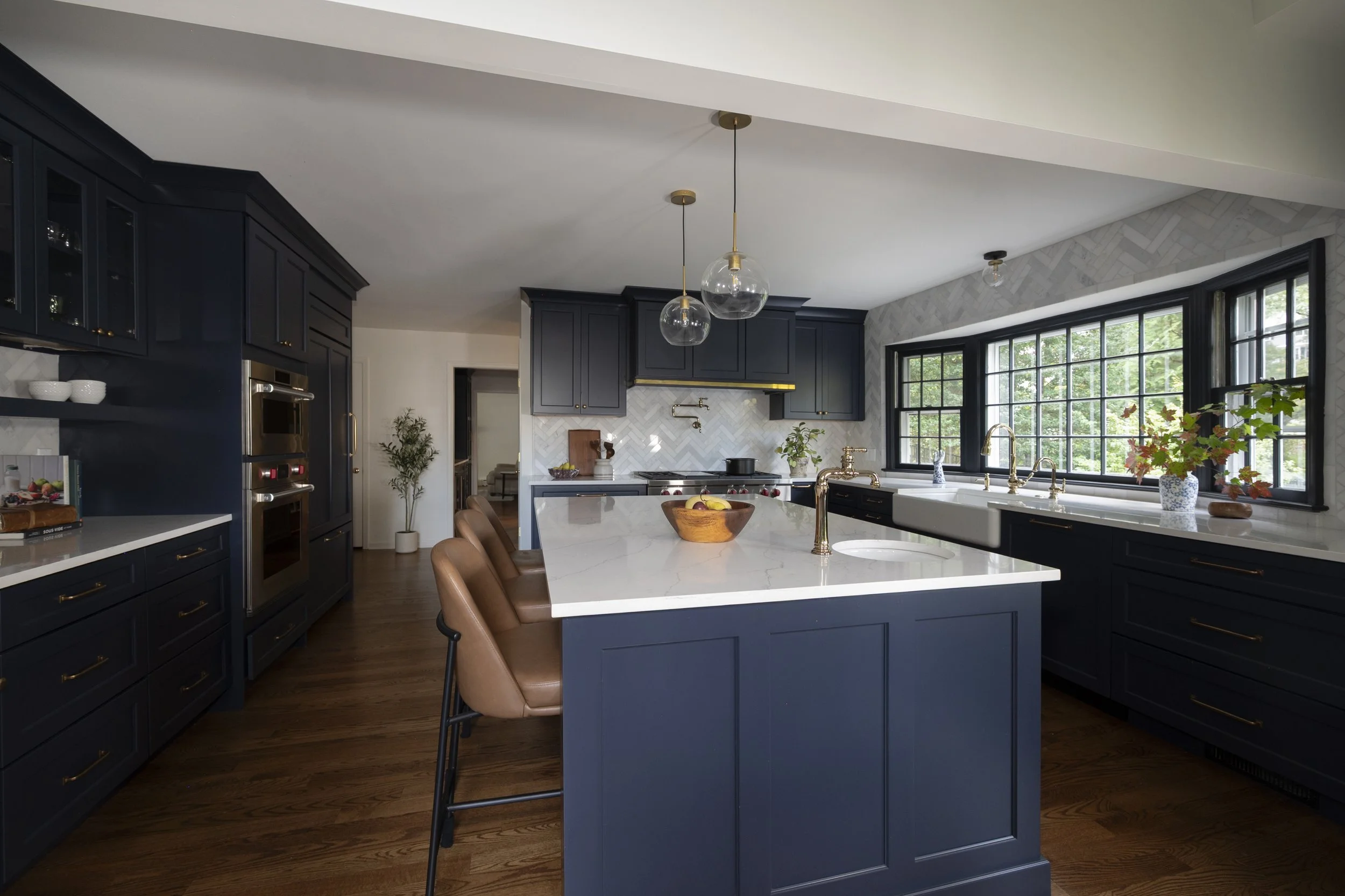 Modern kitchen with navy blue cabinets, a white marble island, large black-framed windows, brass fixtures, and pendant lights. Decor includes a fruit bowl, potted plants, and bowls on the shelves.