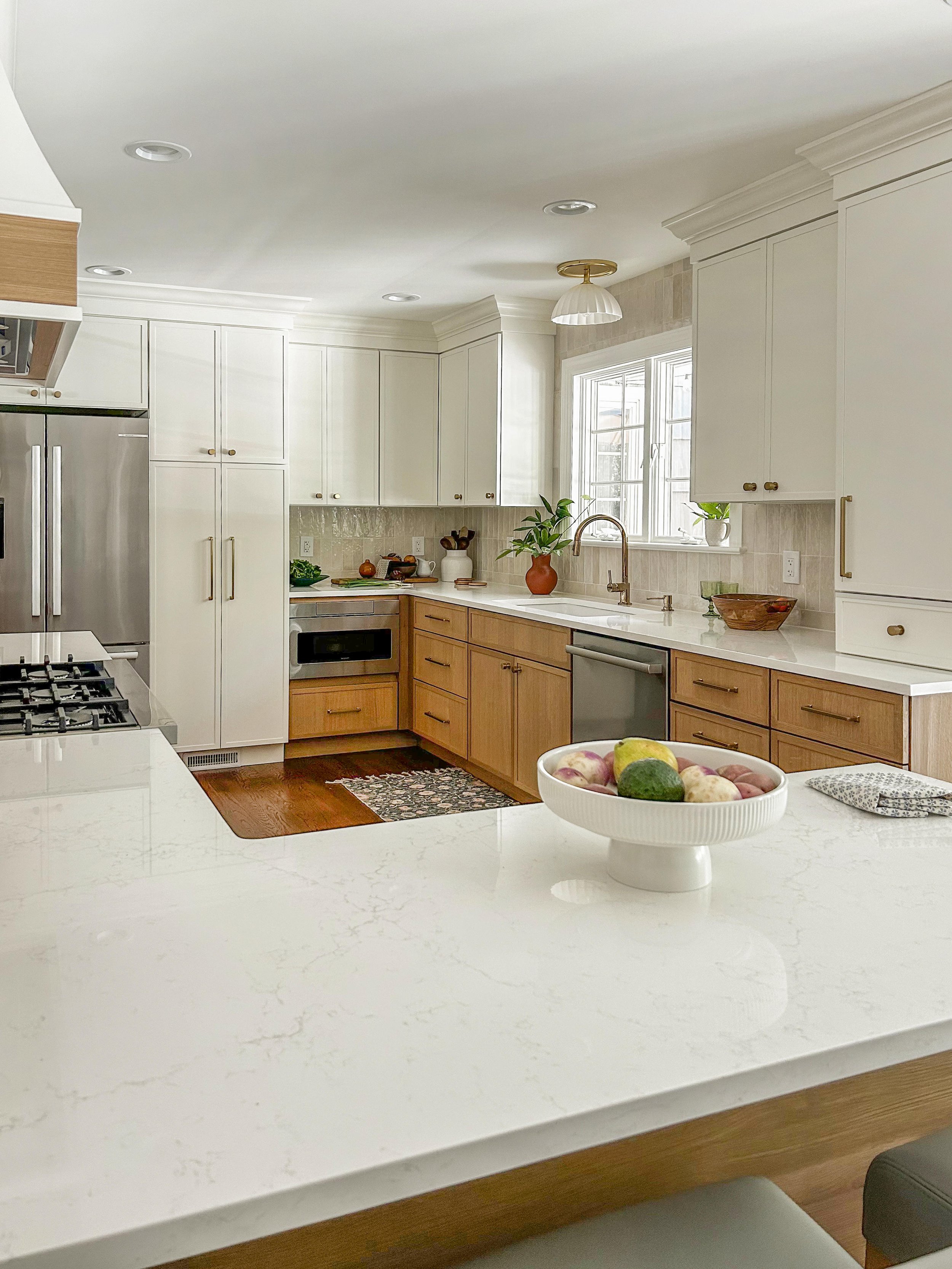 A kitchen with white upper cabinets, wooden lower cabinets, a white marble countertop, a stainless steel dishwasher, a stove, and a window with white trim. There are decorative items and plants on the counter, and a bowl of fruit in the foreground.