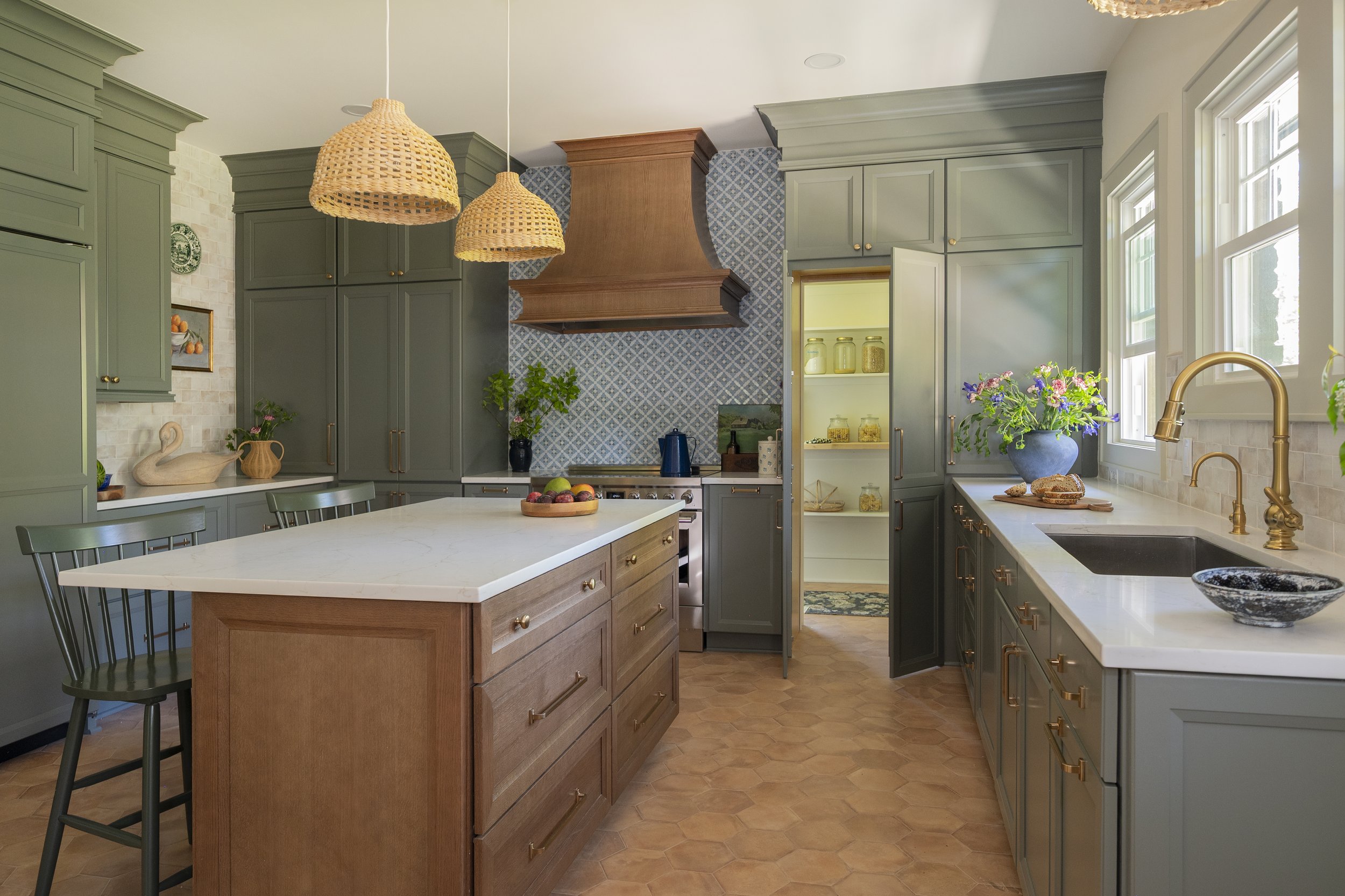 A bright modern kitchen with green cabinetry, white marble countertops, and a large island with wooden drawers. There are woven pendant lights hanging from the ceiling, and a window with a view outside. Decor includes flowers, a bowl of fruit, and jars of preserved food.