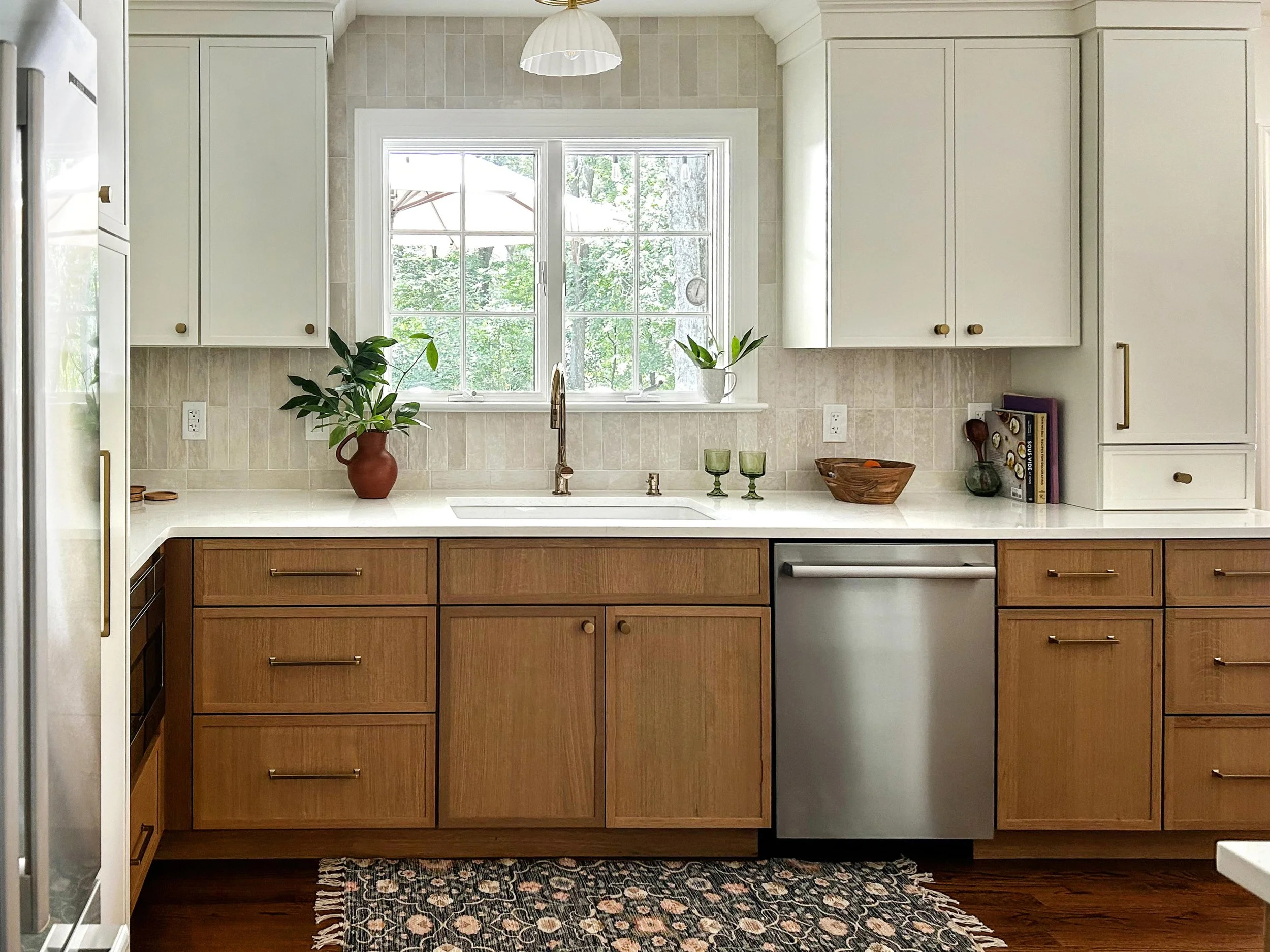 Kitchen with wooden cabinets, a window with a view of trees, a plant, and a rug on a hardwood floor.