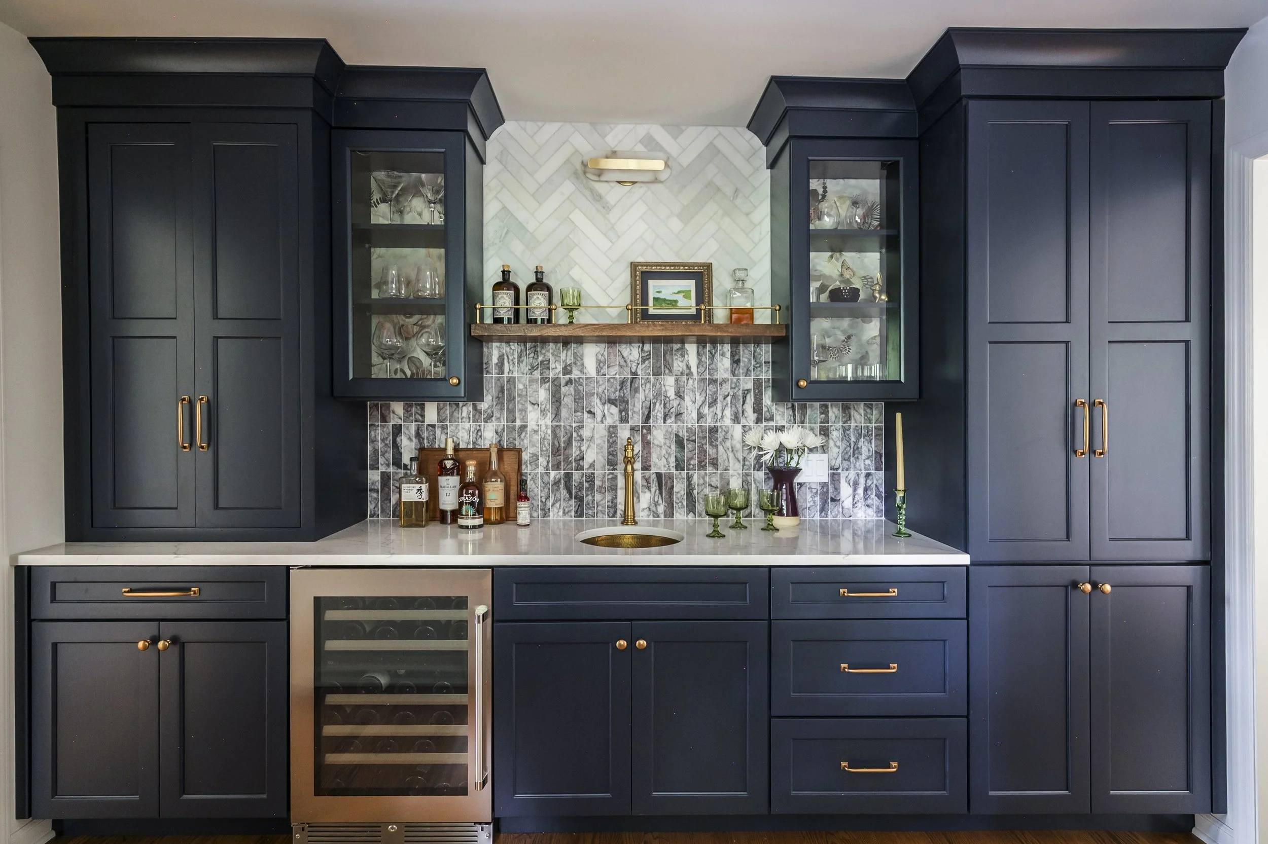 A modern kitchen with dark blue cabinets, a marble backsplash, and a white countertop. There is a small wine fridge, glassware, and decorative items on the countertop and shelves.