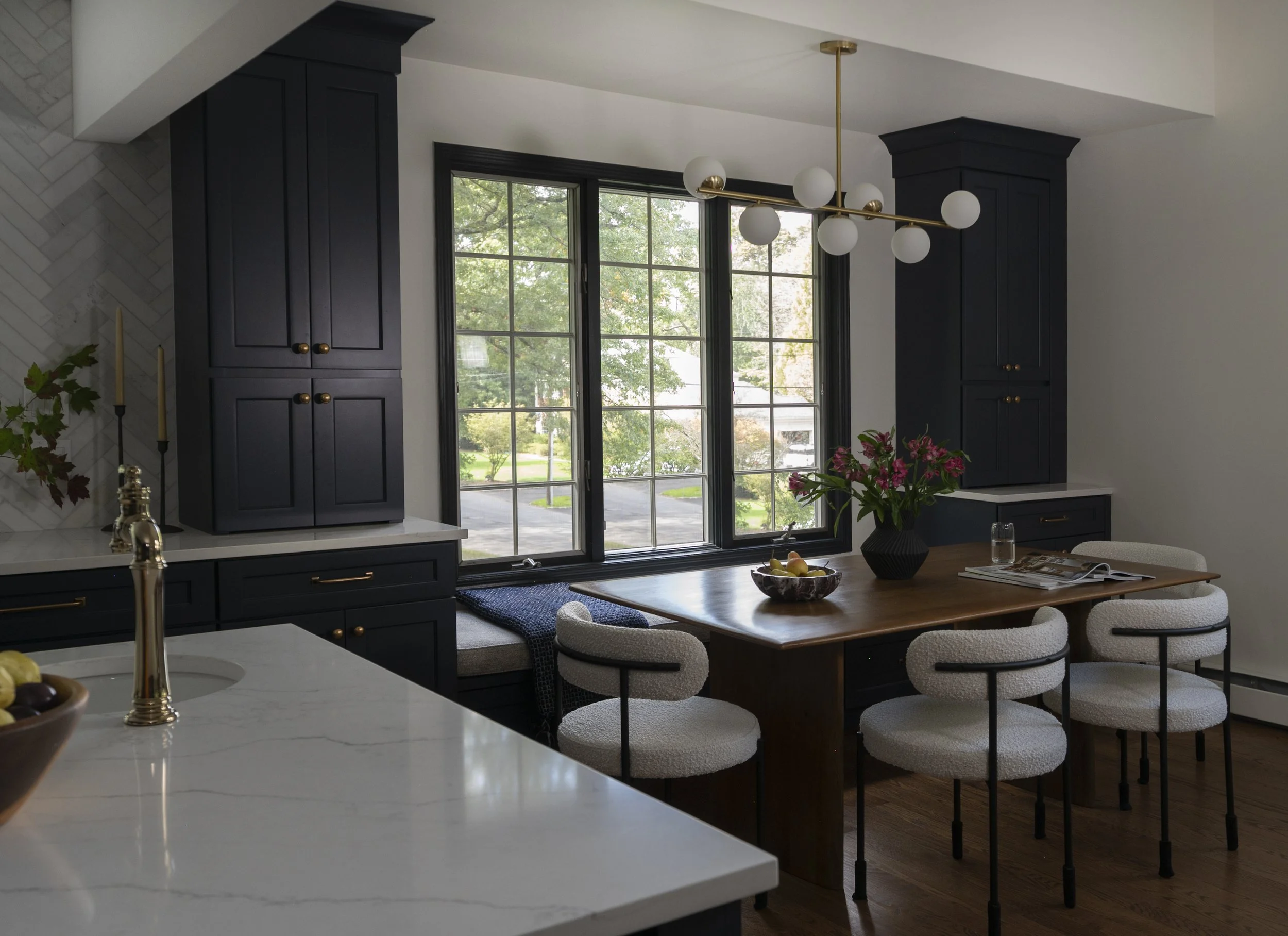 Inside view of a modern kitchen with dark cabinets, large windows, and a wooden dining table with chairs. There is a contemporary chandelier hanging from the ceiling and a vase with pink flowers on the table.