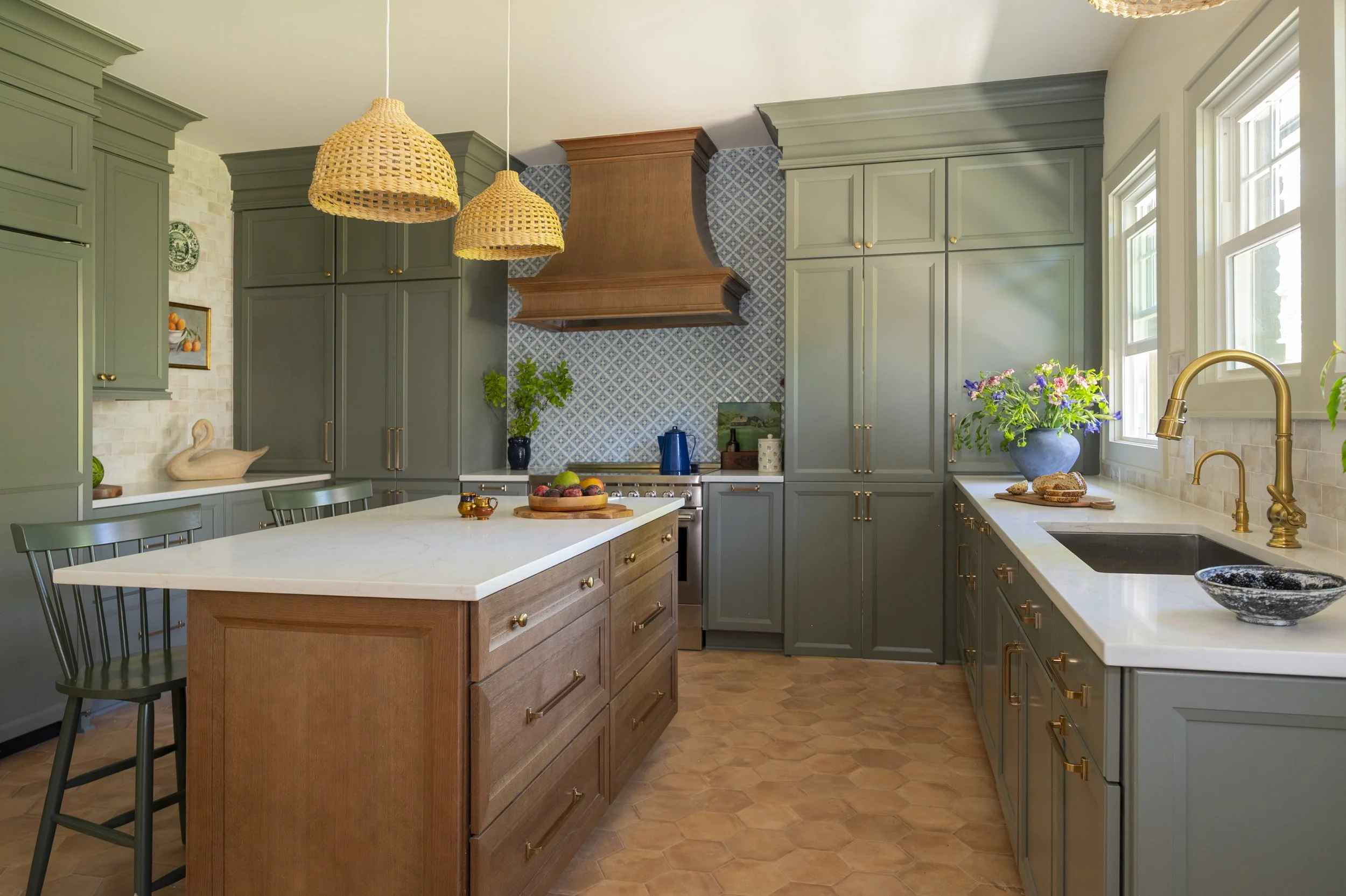 A kitchen with green cabinets, a wooden island, a white countertop, and a hexagonal tile floor. There are two woven pendant lights hanging from the ceiling, a window with a flower vase on the counter, and a stove with a wooden range hood against a blue patterned backsplash.