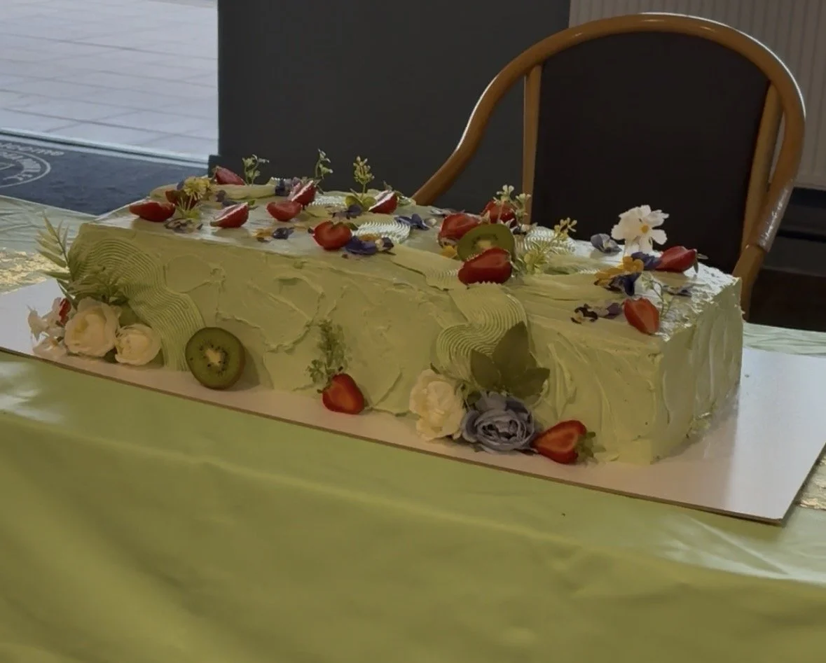 A rectangular cake decorated with green frosting, topped with strawberries, blueberries, and various edible flowers, placed on a white board on a green tablecloth.