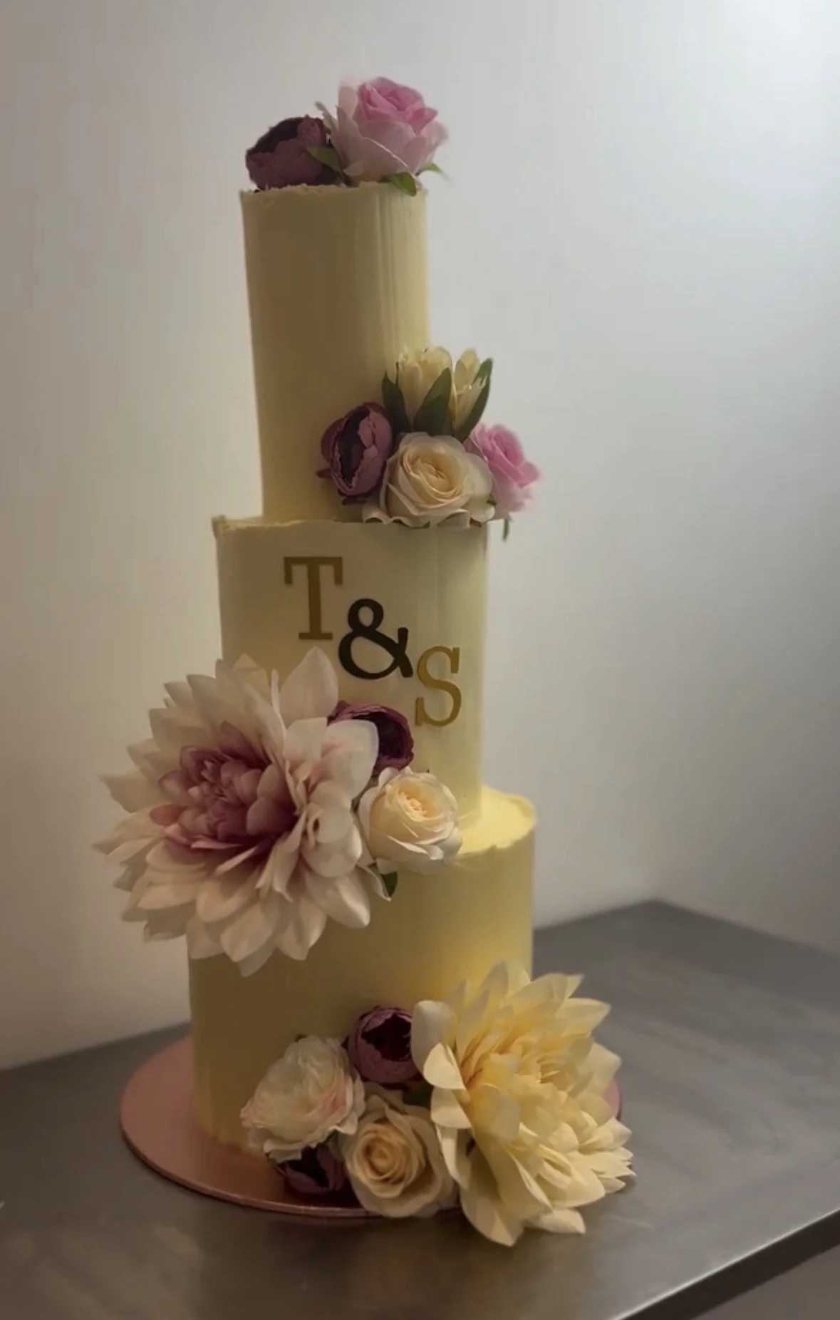 A tiered wedding cake decorated with pink, white, and purple flowers, with the initials T&S on the middle tier.