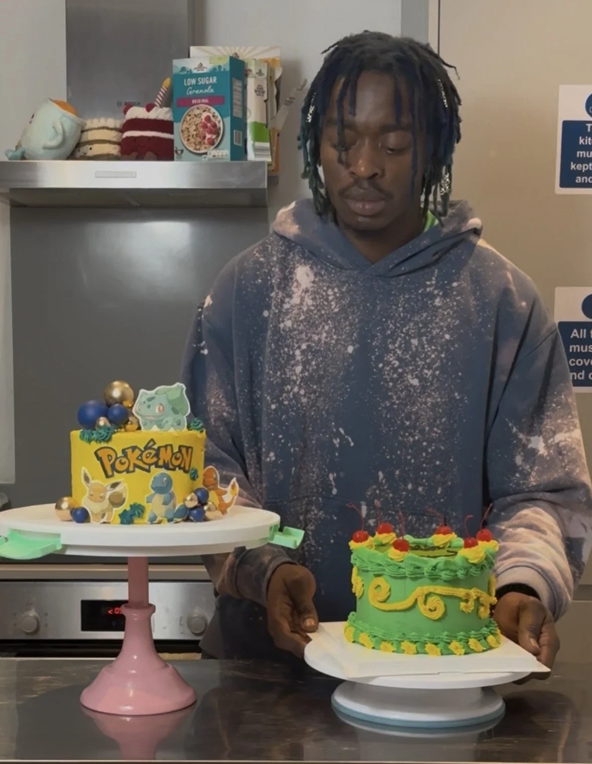 Zhora wearing a gray hoodie with white paint splatters holds two decorated cakes, one with Pokémon characters and the other with yellow and green swirls and cherries, in a kitchen with shelves of food and signs on the wall.