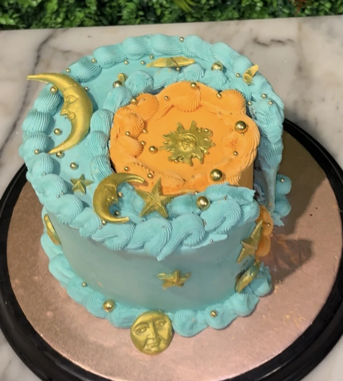 A decorative cake with a celestial theme featuring crescent moons and stars in gold, with blue and orange frosting and gold embellishments.