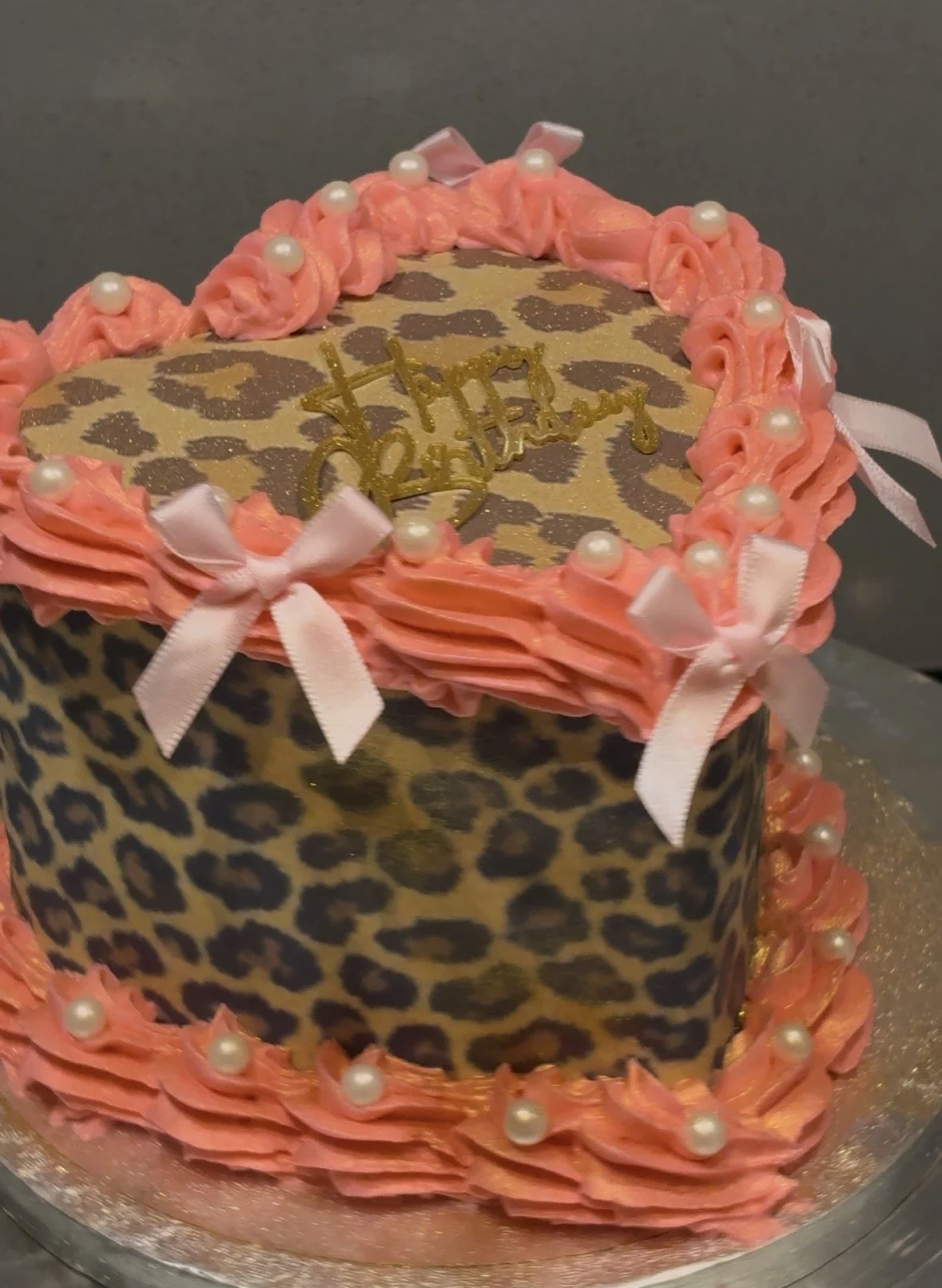 A decorated cake with leopard print pattern, pink frosting, pearl-like accents, and pink bows, with a gold 'Happy Birthday' topper.