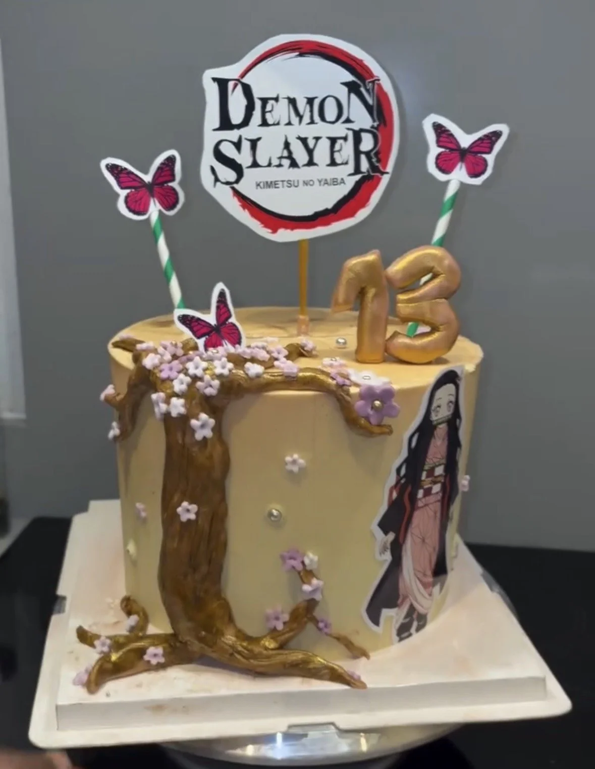 A birthday cake decorated with "Demon Slayer" theme, featuring a "Demon Slayer" logo topper, pink butterfly decorations, a character cutout, pink flowers, and the number 13 in golden frosting.