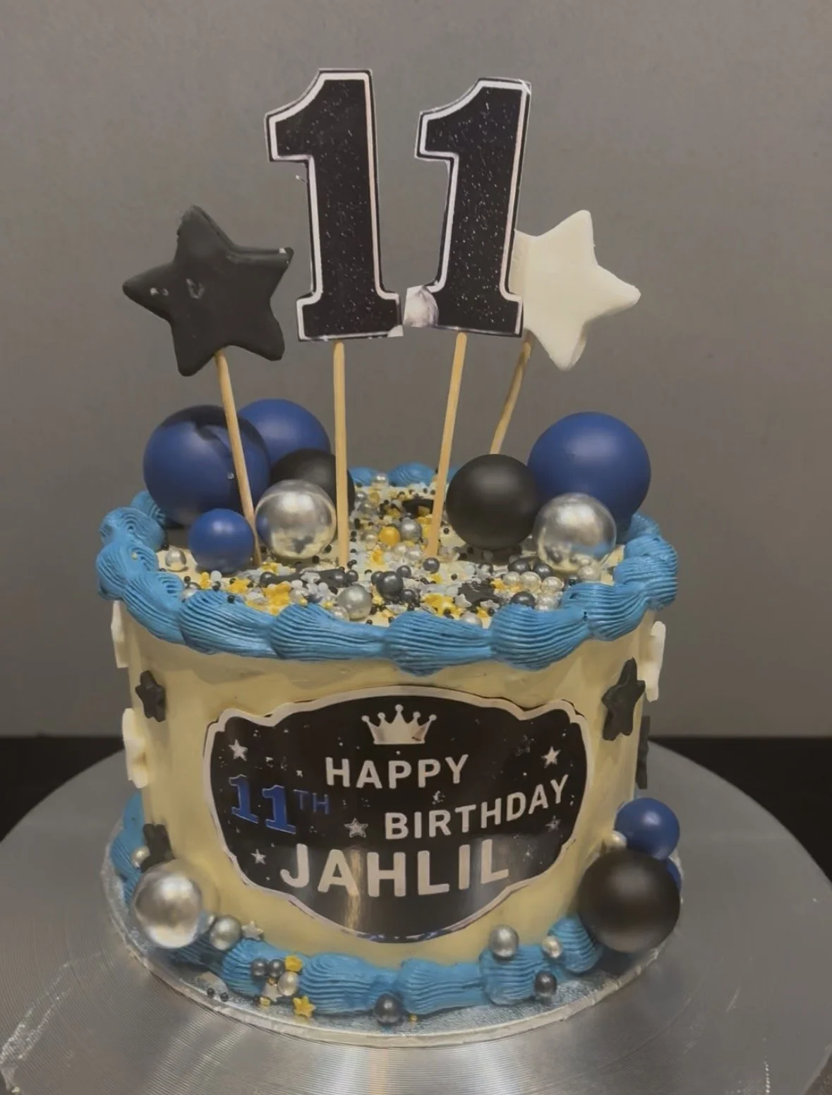 A birthday cake decorated with blue, black, and silver spheres, stars, and a topper with the number 11 and stars, celebrating an 11th birthday for Jahil.