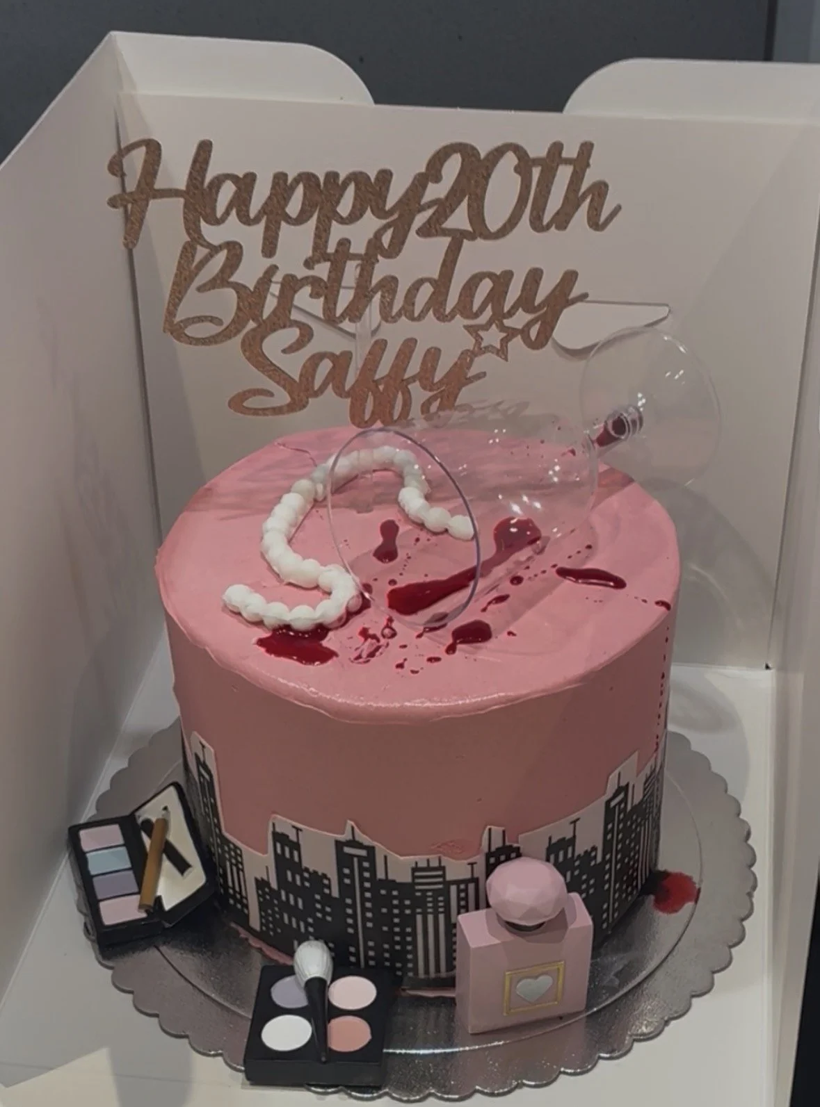 Pink birthday cake with cityscape design, topped with clear balloons, an 'Happy 20th Birthday Saucy' sign, and various makeup-themed decorations and tools surrounding it.