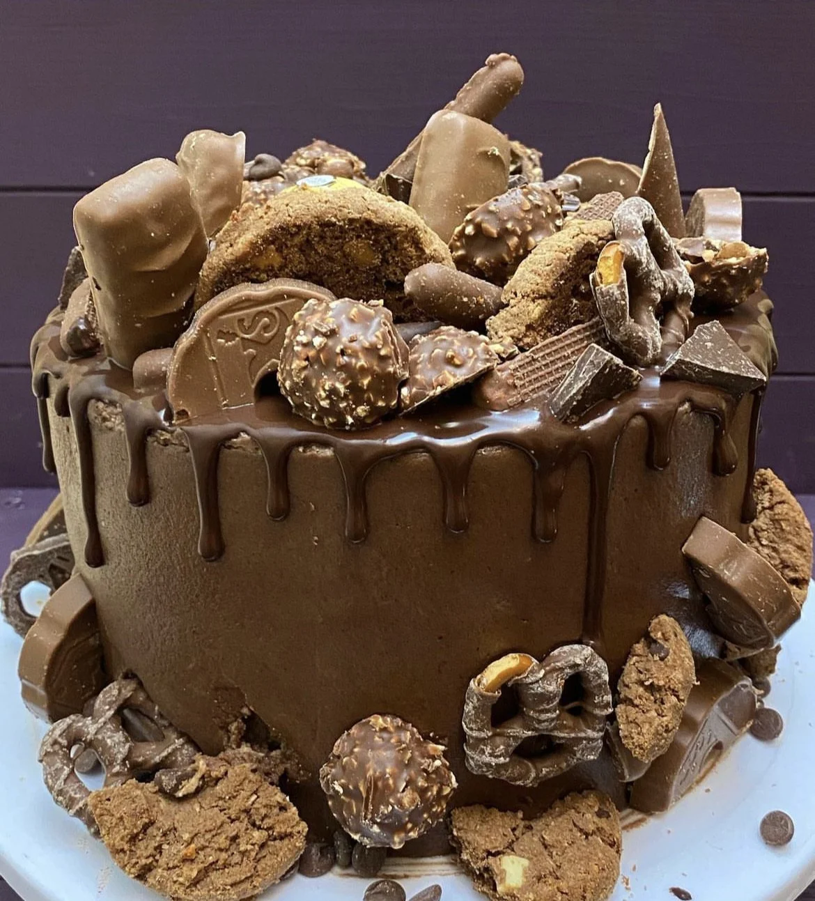 A large chocolate cake topped with various chocolates, cookies, and chocolate-covered candies, with a chocolate ganache drizzle on top.
