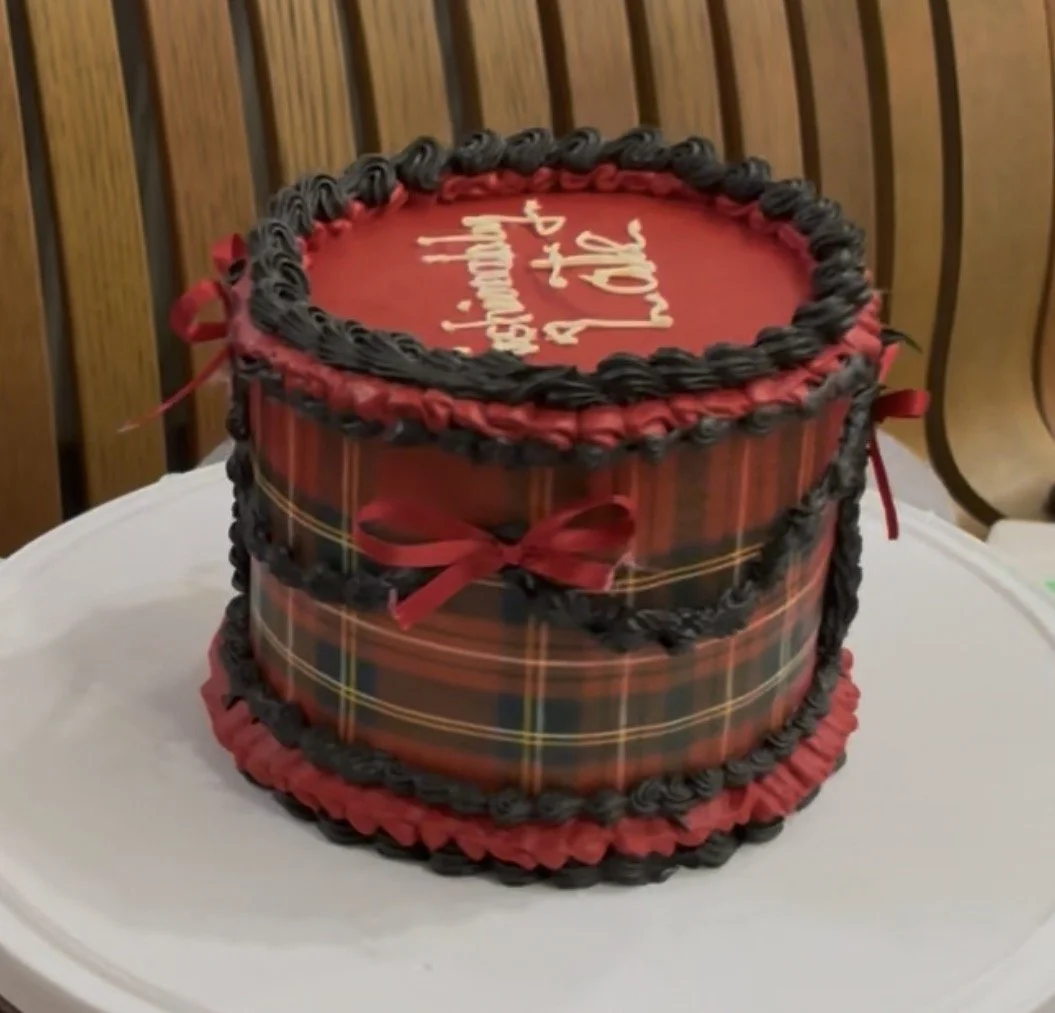 A round birthday cake with red and black tartan plaid decor, black piped border on top and bottom, red ribbon accents, and a message written on top in white icing.