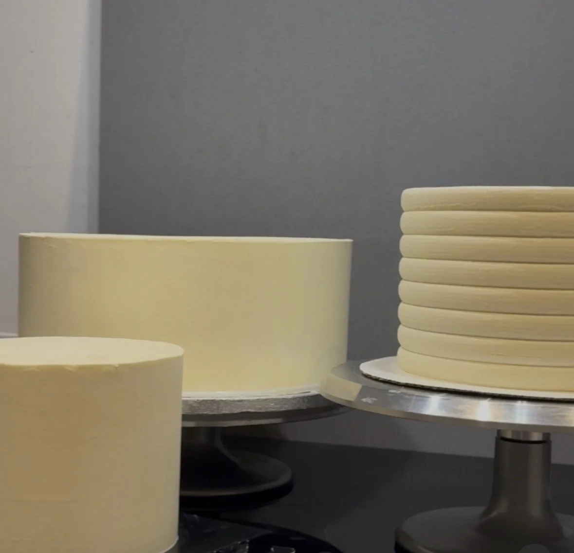 Multiple plain, uniced cake layers stacked on metal cake stands against a grey wall.