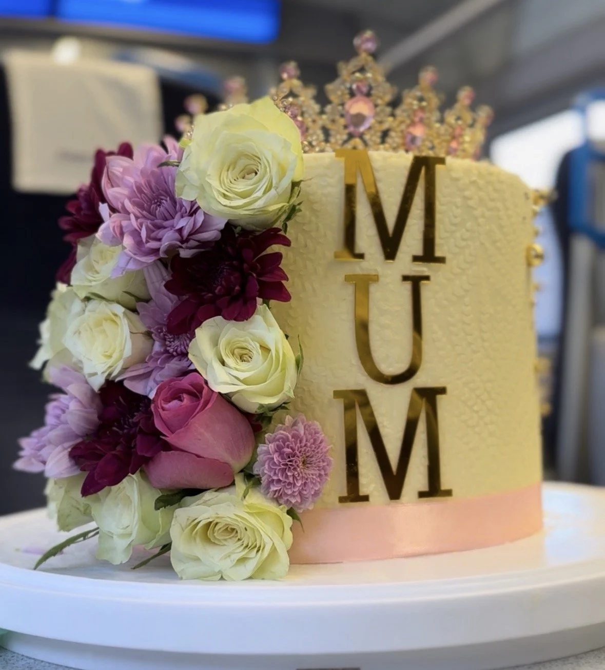 A decorated cake with white and pink flowers on the side, topped with a crown, and gold letters that spell 'MUM' in the center.