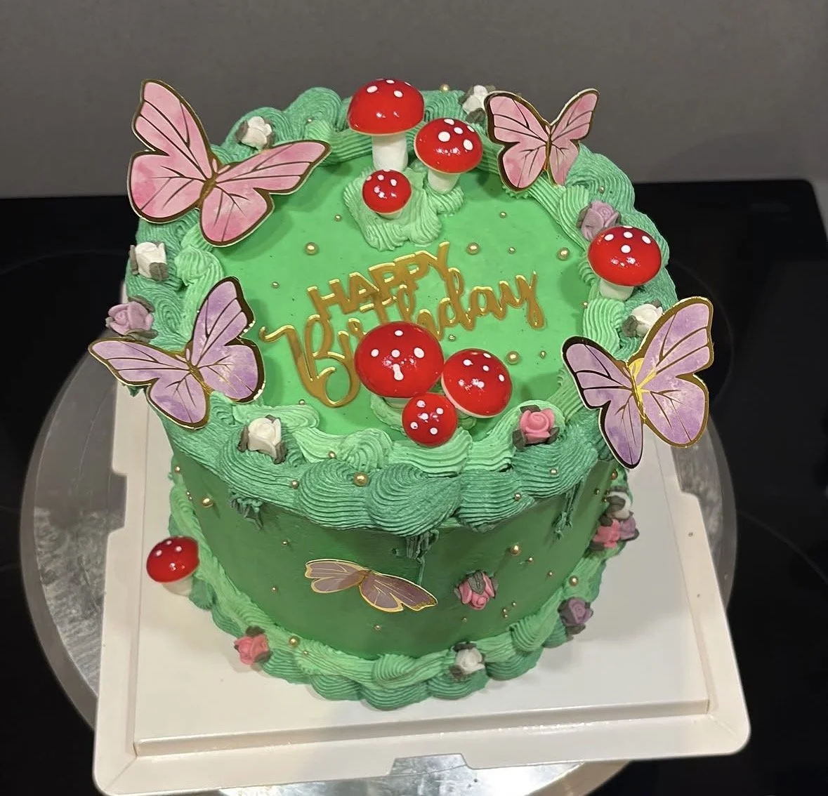 A green birthday cake decorated with red and white mushrooms, pink and purple butterflies, small pink and white flowers, and golden balls. The top of the cake has a gold 