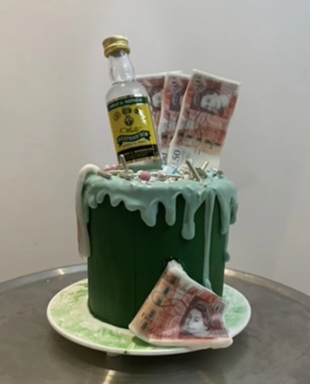 A green birthday cake decorated with white icing, topped with a small bottle, banknotes, beads, and sprinkles, with a banknote placed on a plate in front of the cake.