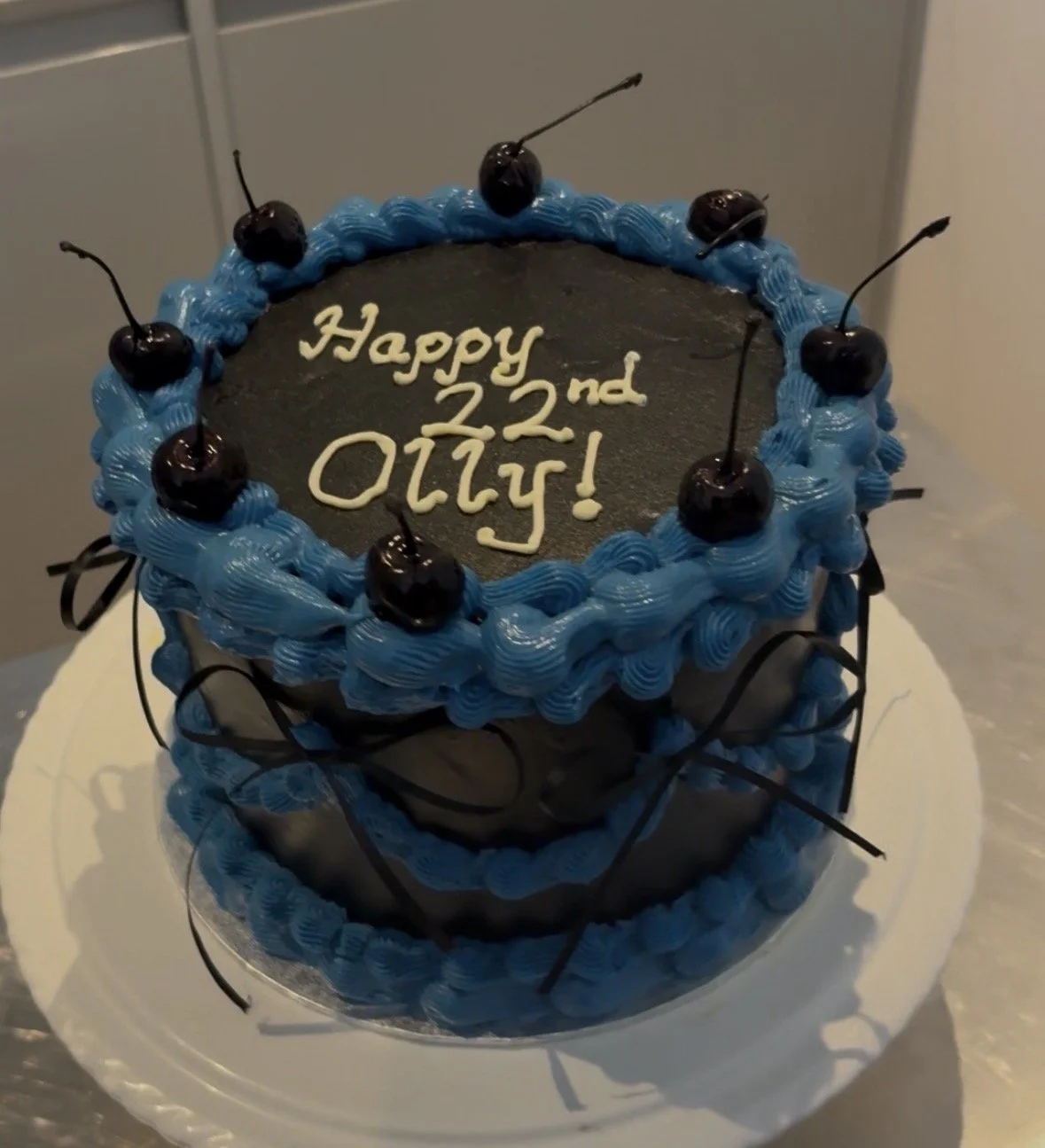Birthday cake with blue frosting, chocolate cherries, and black decorative curls, with a message that reads, 'Happy 22nd Ollie!'