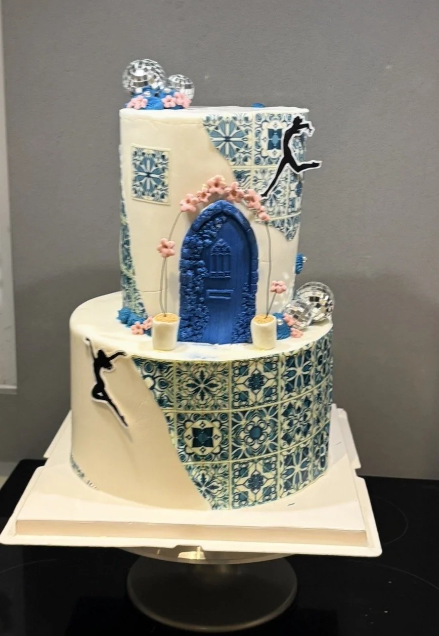A two-tiered cake decorated with blue and white tiles, featuring dancing silhouettes and disco balls, celebrating a dance or ballet theme.