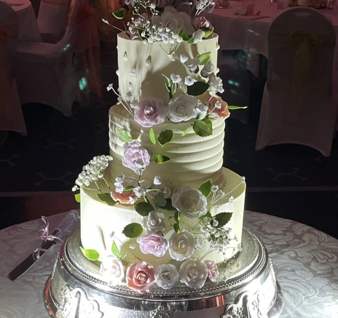 Three-tiered wedding cake with white icing decorated with pink and white roses, green leaves, and small white flowers, sitting on a silver cake stand.