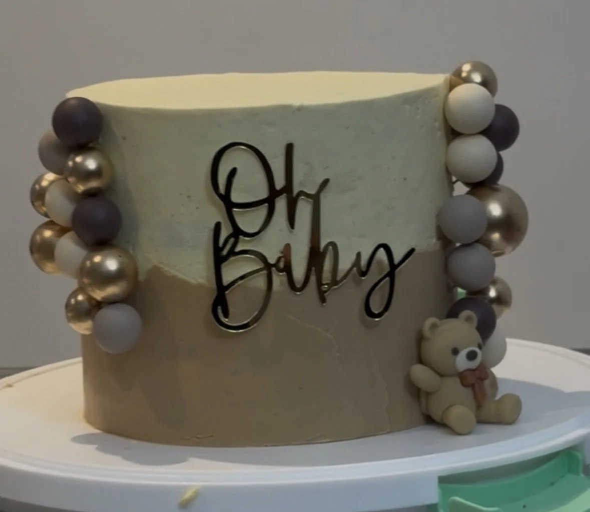 A cake with a beige and white marble pattern, decorated with silver, gray, and white edible beads on the sides, a small teddy bear figure at the base, and a black script "Oh Baby" topper.