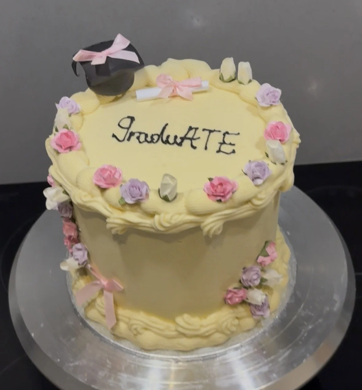 A round cake decorated with pink, purple, and white flowers and a pink bow, with the word 'Graduation' written on top, topped with a black graduation cap with a pink ribbon.