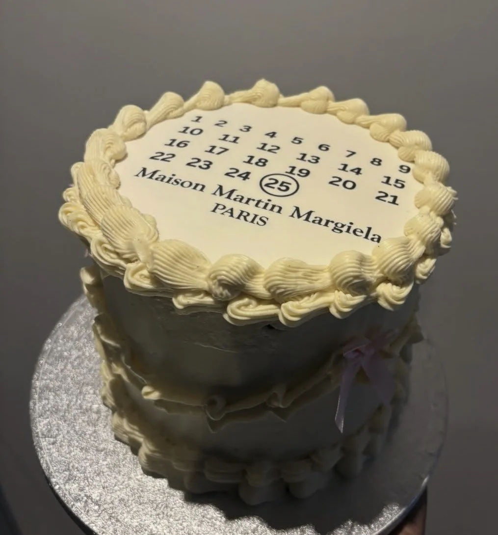 A two-tier cake decorated with white frosting and piped borders, featuring a design inspired by a calendar with numbers from 1 to 25, and the names "Maison Martin Margiela PARIS."