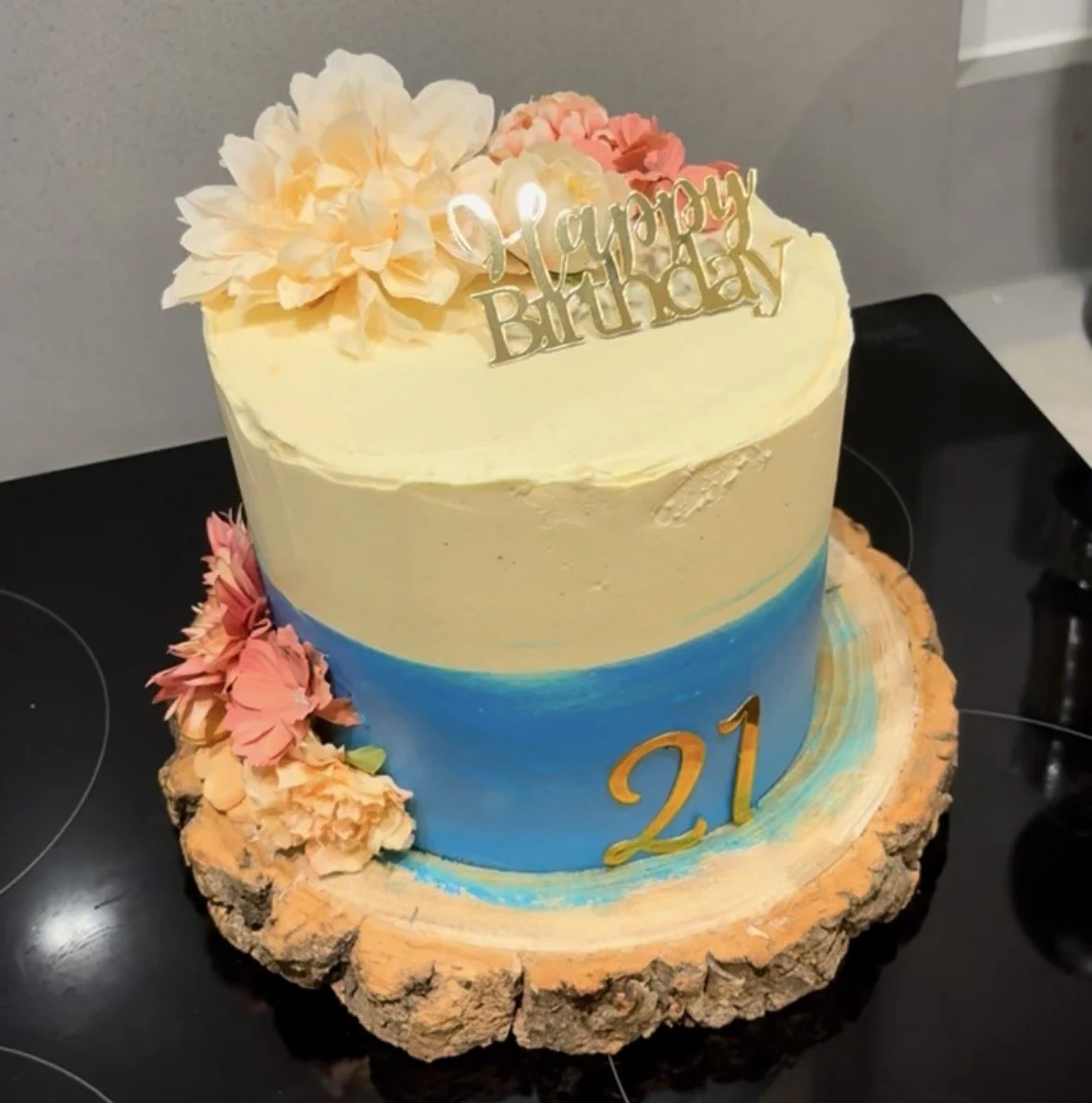 A two-tier birthday cake with a blue bottom tier and a cream top tier, decorated with pink and yellow flowers, a "Happy Birthday" topper, and the number "21" on the cake board. The cake is on a wooden slab.