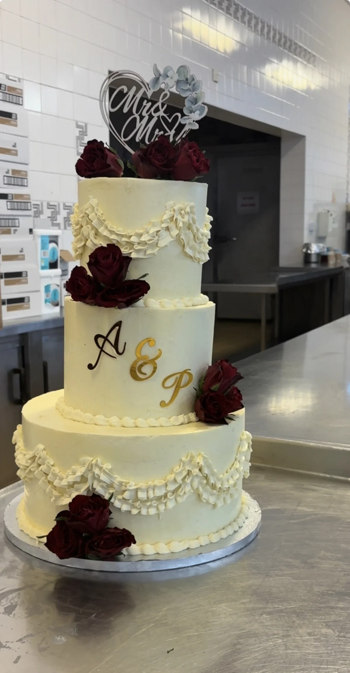 Three-tier wedding cake decorated with white icing, red roses, and white piped details, with a "Mrs & Mrs" topper featuring flowers, on a metal table in a kitchen or bakery setting.