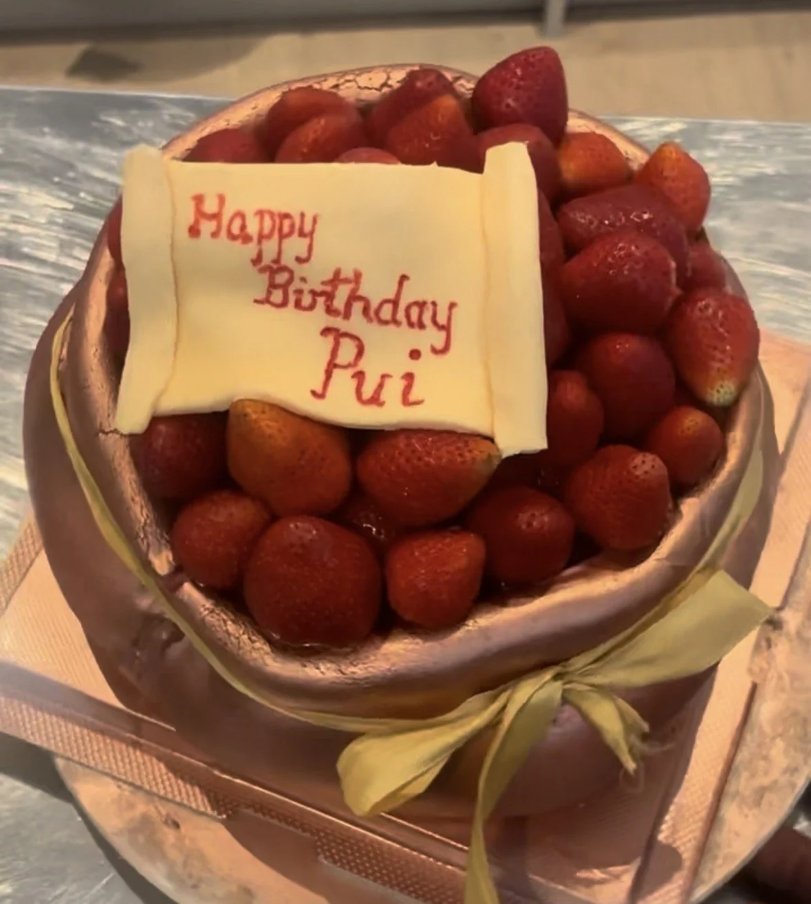 A round birthday cake decorated with strawberries and a white chocolate plaque with red writing that says, 'Happy Birthday Pui'.